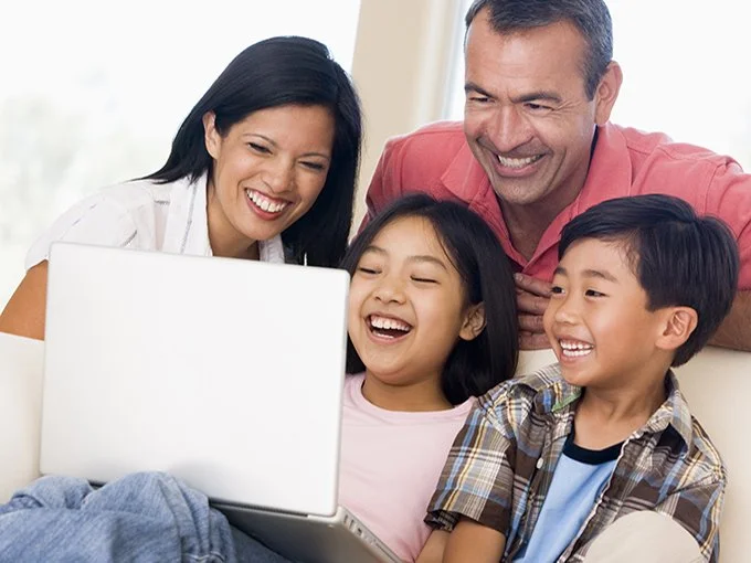 Happy family of four sitting on couch, laughing together while looking at a laptop screen.