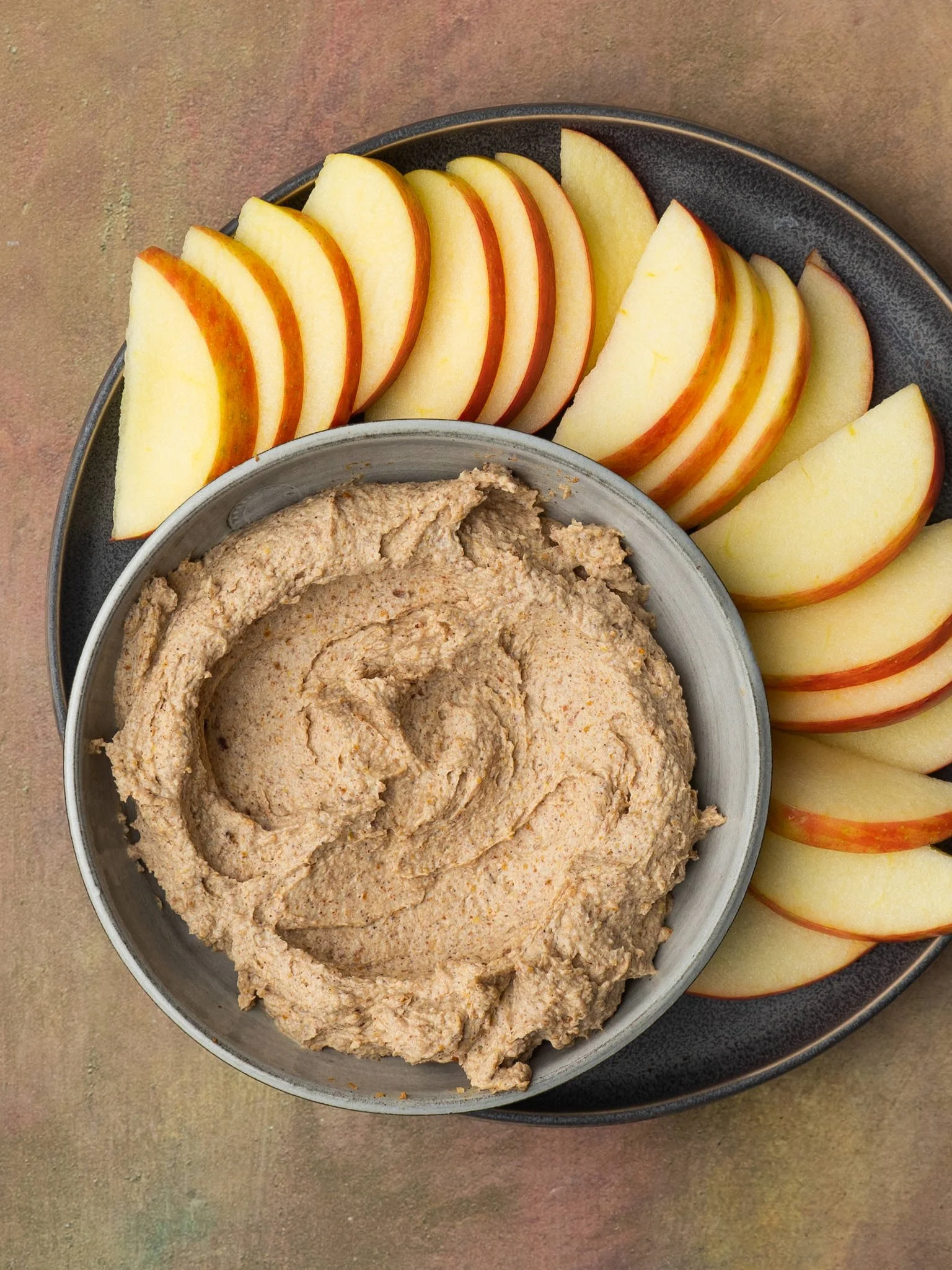 A bowl of creamy almond butter sits in the center of a plate surrounded by neatly arranged fresh apple slices.