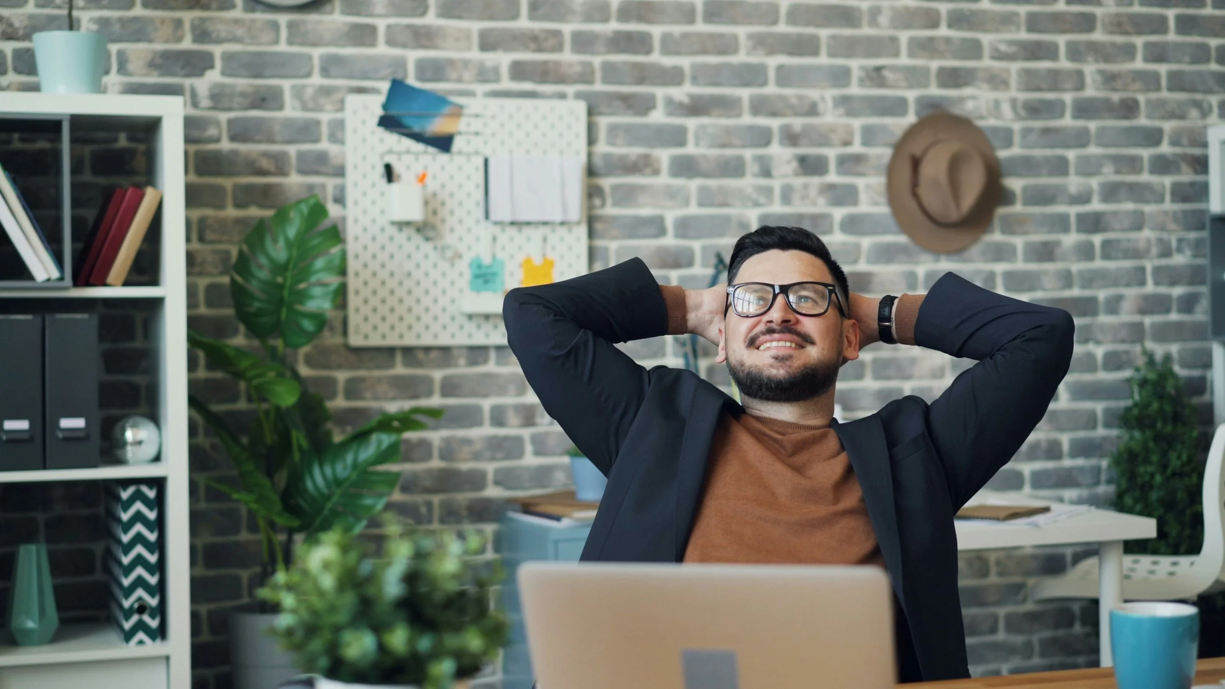 Man in glasses leaning back in chair and smiling at his desk, appearing relaxed in a modern home office.