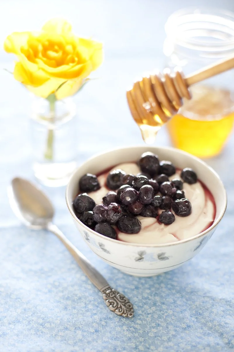 Honey is being drizzled over a bowl of yogurt topped with blueberries, next to a yellow rose and a jar of honey on a table.