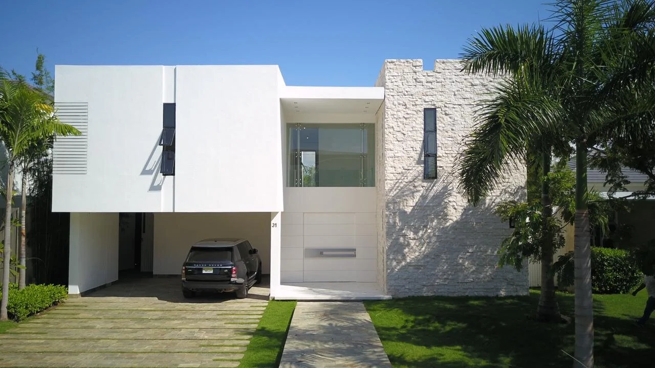 Modern two-story house with white and stone exterior, palm trees, a driveway with a black SUV, and a clear blue sky.