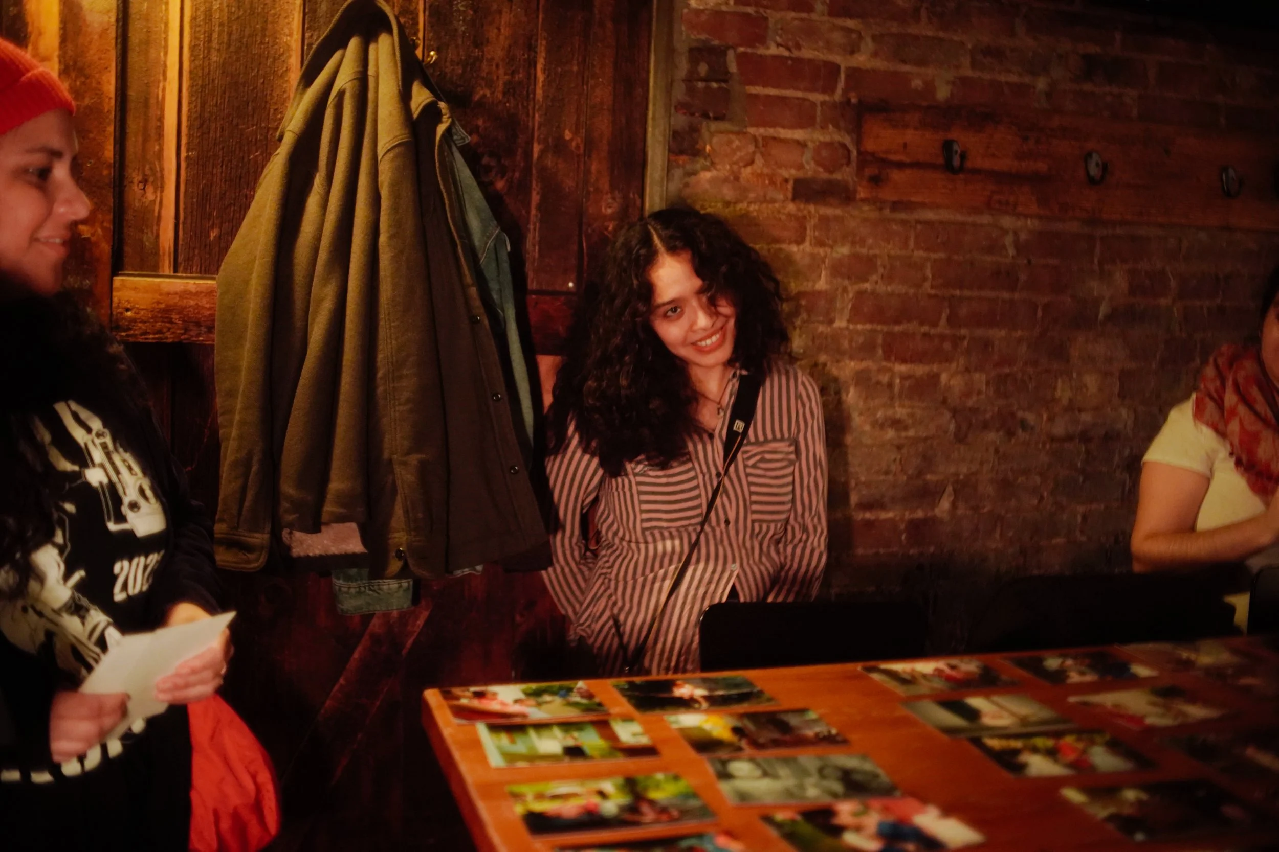 A young woman with dark curly hair wearing a striped shirt, smiling and leaning against a wooden wall in a dimly lit room with a brick wall. There are photos on a table in front of her, and coats hanging on the wall behind her.
