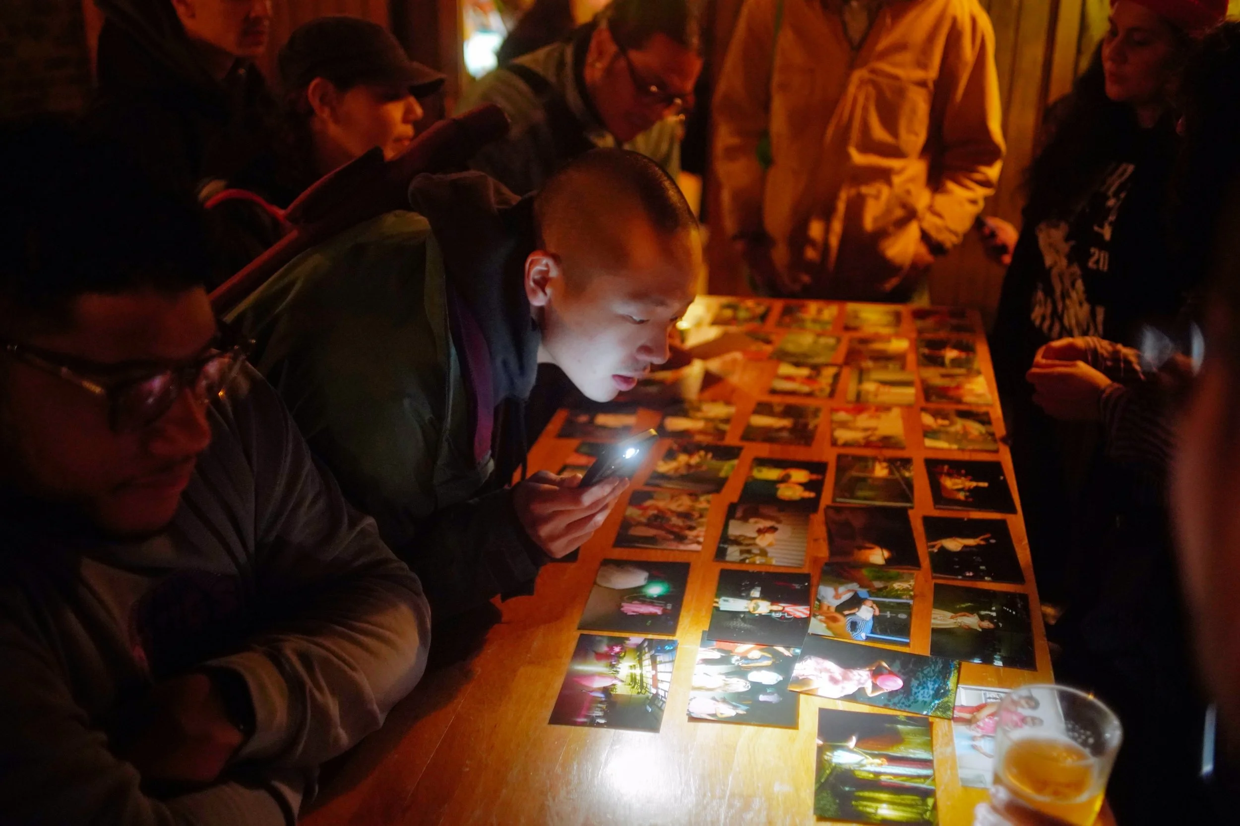 People gathered around a table looking at printed photographs, with one person using a phone flashlight to see clearer. The setting appears to be a dimly lit room, possibly a bar or social event.