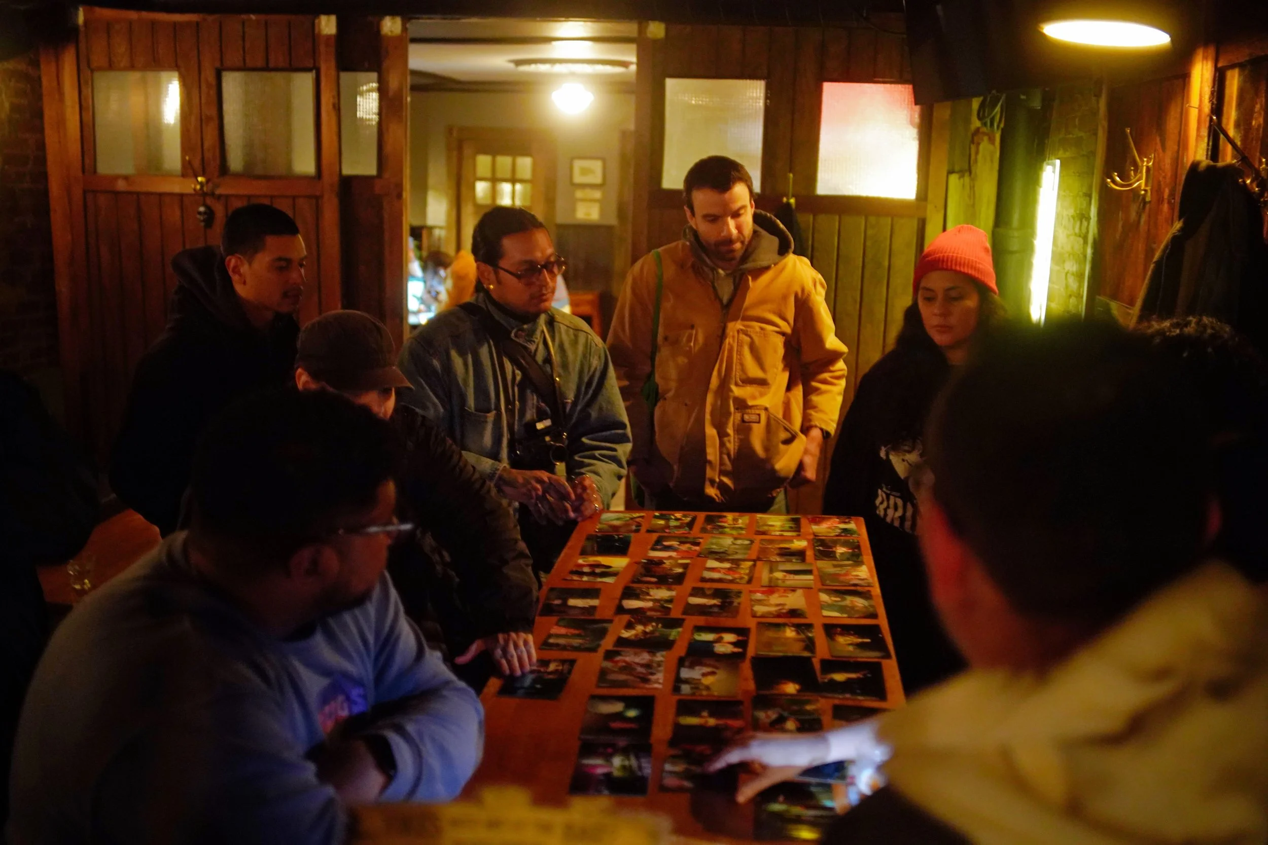 A group of people gathered around a table looking at numerous photographs displayed on a wooden table inside a dimly lit rustic room with wood-paneled walls and warm lighting.