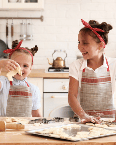 Two young girls with pigtails wearing aprons and red polka-dotted headbands baking cookies in a kitchen from their snail mail for kids club.