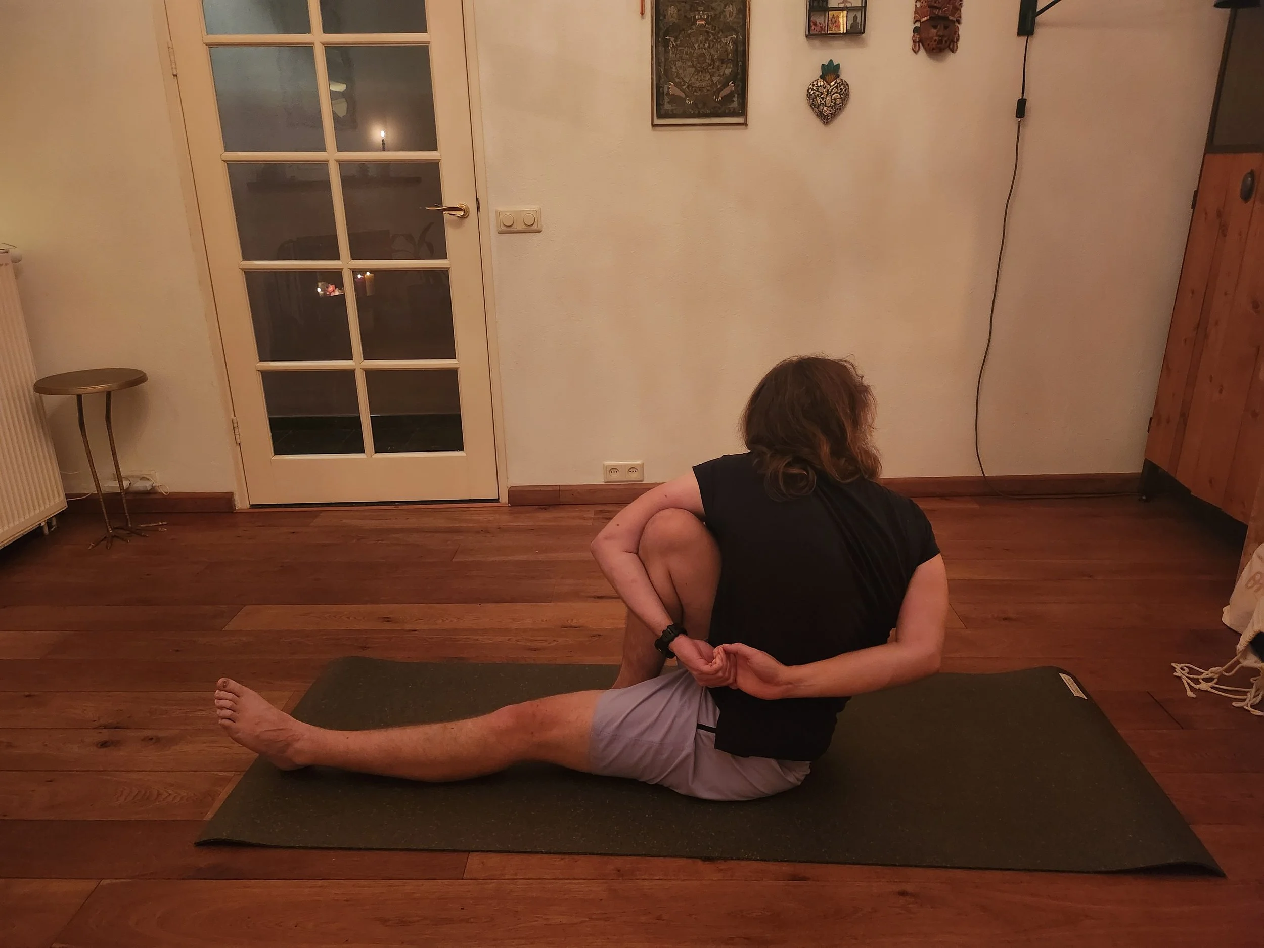 A person practicing yoga indoors on a black mat, sitting on one side with one leg extended forward, the other leg bent and held behind them with one hand, and the other arm reaching back to grasp the foot of the bent leg. The room has a wooden floor, a glass-paneled door, and wall decorations.