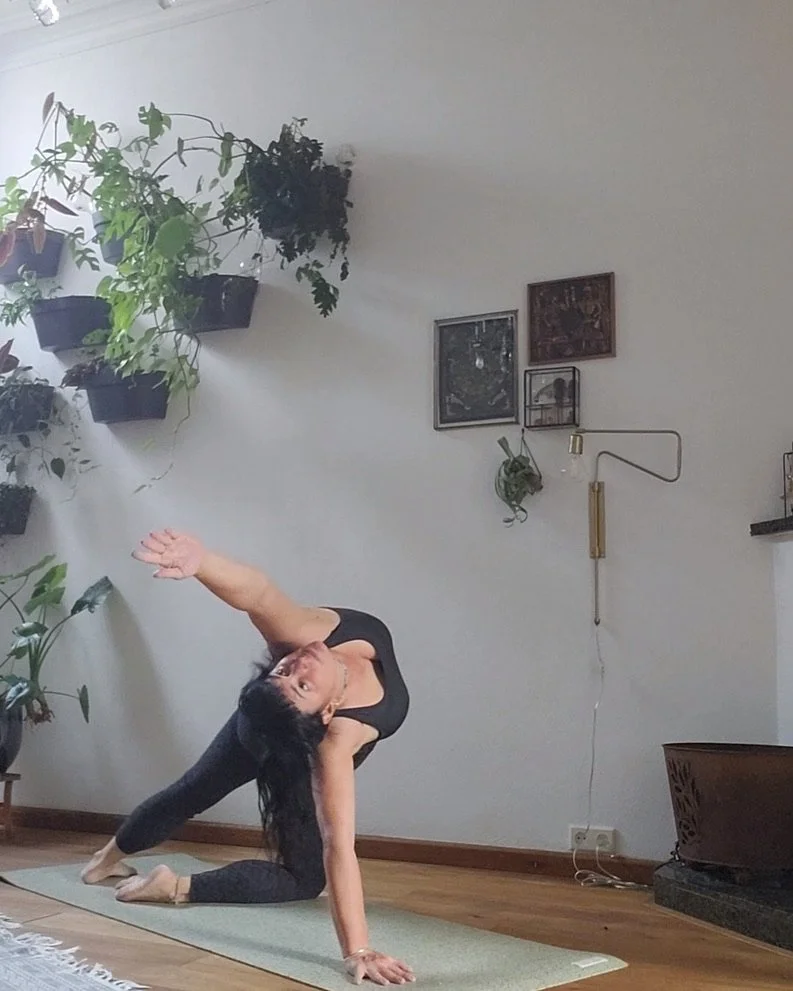 A woman practicing yoga in a room with potted plants on the wall and framed pictures, on a green yoga mat.