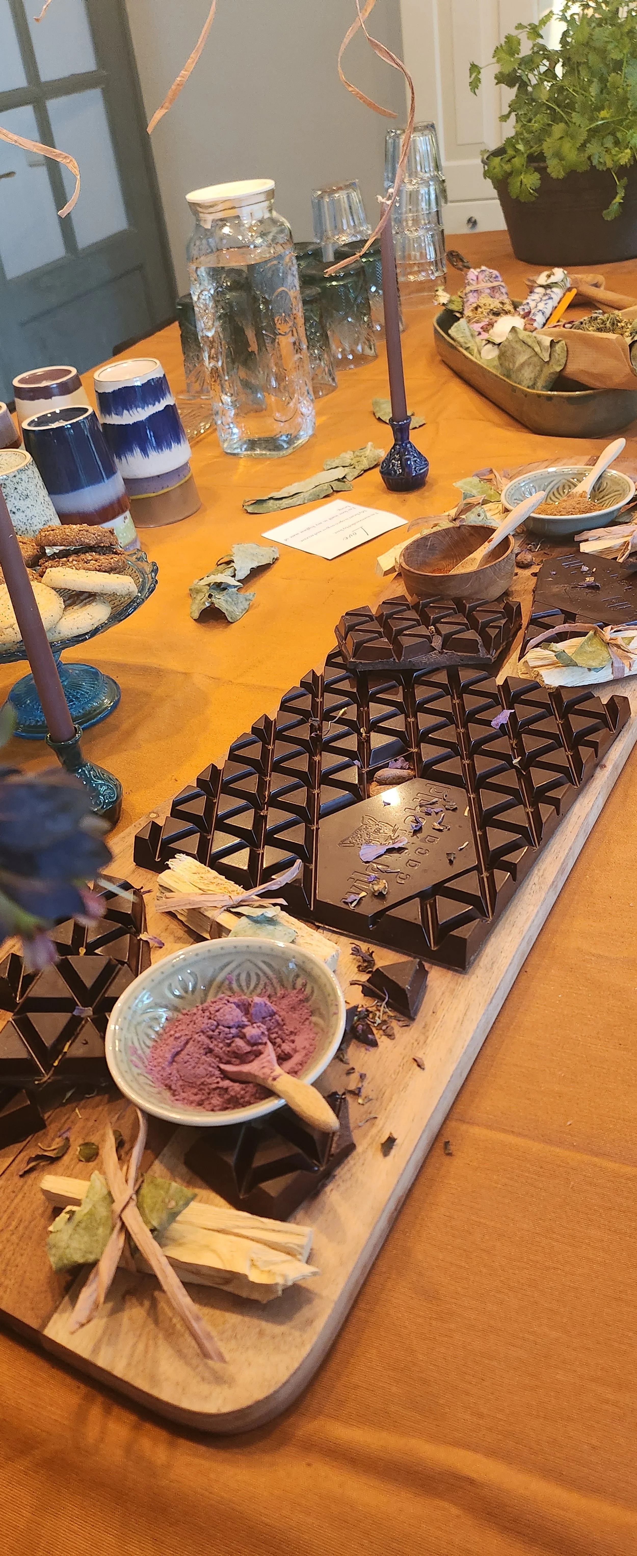 Decorative table with dark chocolate, purple powder in a bowl, wooden board, candles, and assorted tableware.