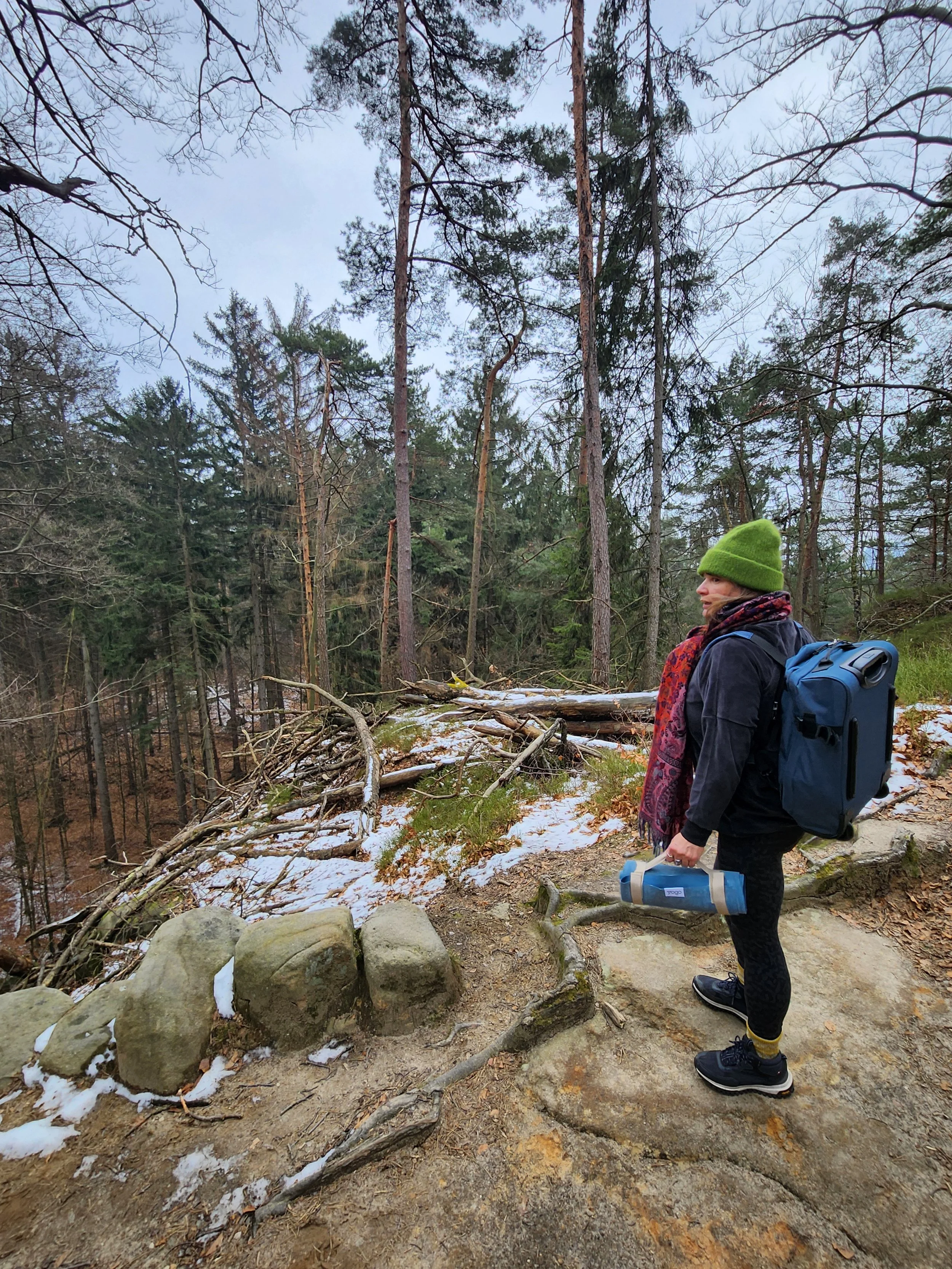 A woman standing on a dirt trail in a forest, holding a rolled-up mat in her right hand and wearing a green beanie, black jacket, patterned scarf, black leggings, and hiking shoes. She has a large backpack on her back.