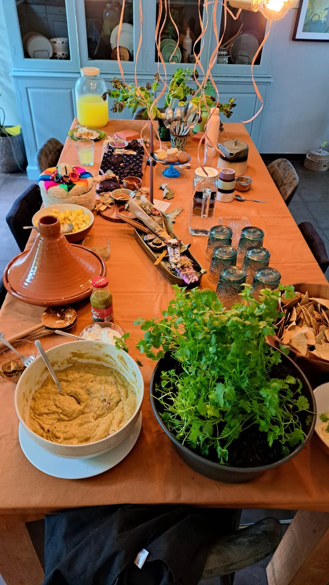 Table set with assorted foods and decorations, including a bowl of dip, a clay tagine, herbs, snacks, and colorful decorations. Glasses and utensils are also visible on the table.
