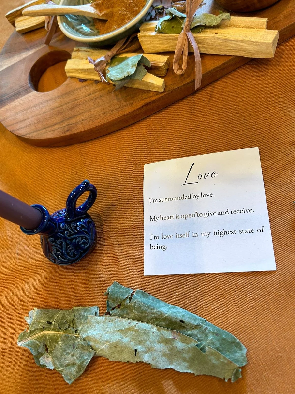 Close-up of a wooden board with Palo Santo sticks tied with ribbons, a small bowl of brown powder with a wooden spoon, dried leaves, and a ceramic candle holder on a table. A card with a message about love is partially visible.