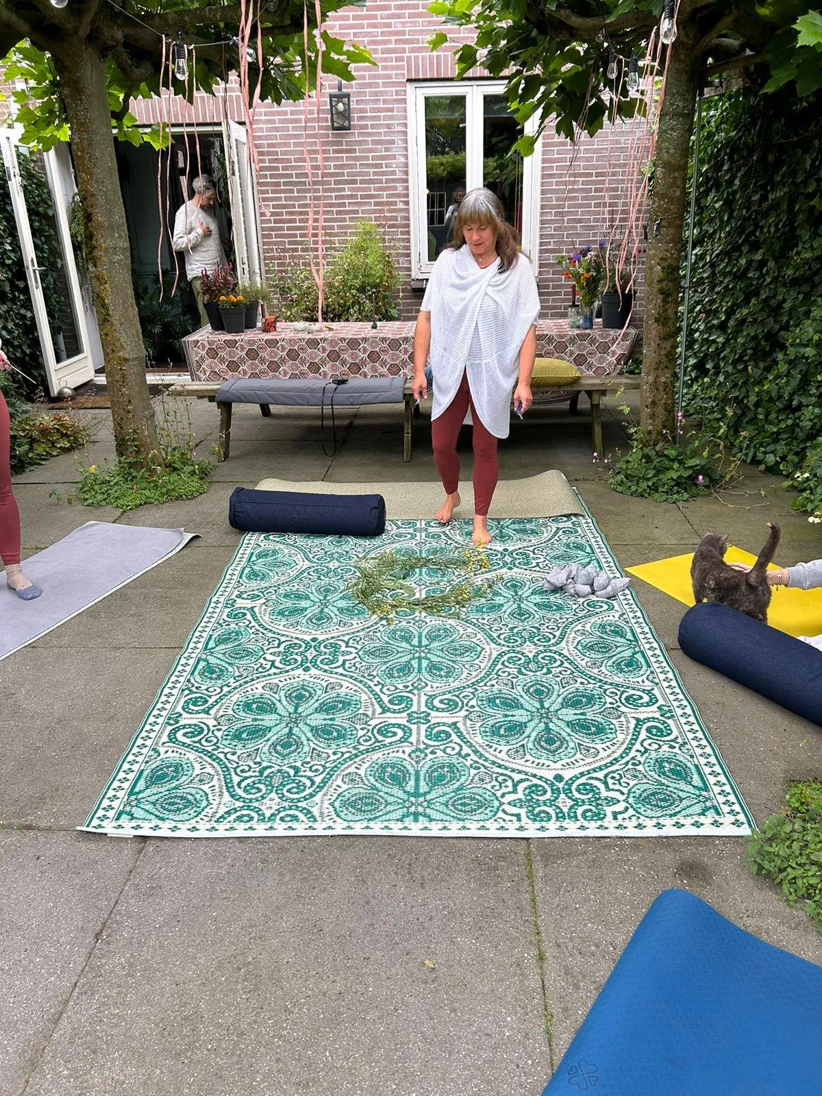 Outdoor yoga setup with decorative green mat, exercise mats, and a woman standing on patio next to a dog.