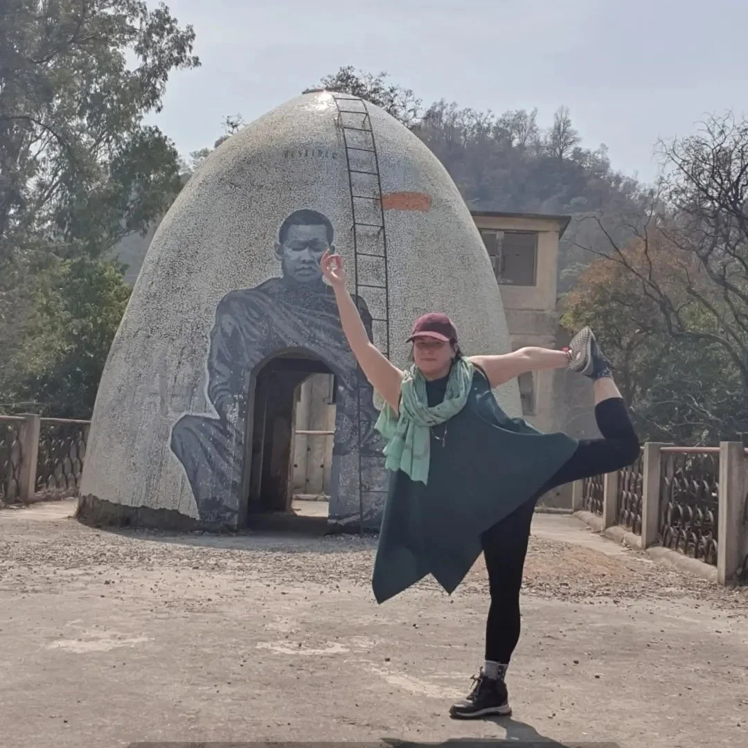 A woman performing a dance pose in front of a small domed building with a mural of a person painted on it. She is balancing on one leg with her other leg lifted behind her, and her arms extended, one upward and one outward.