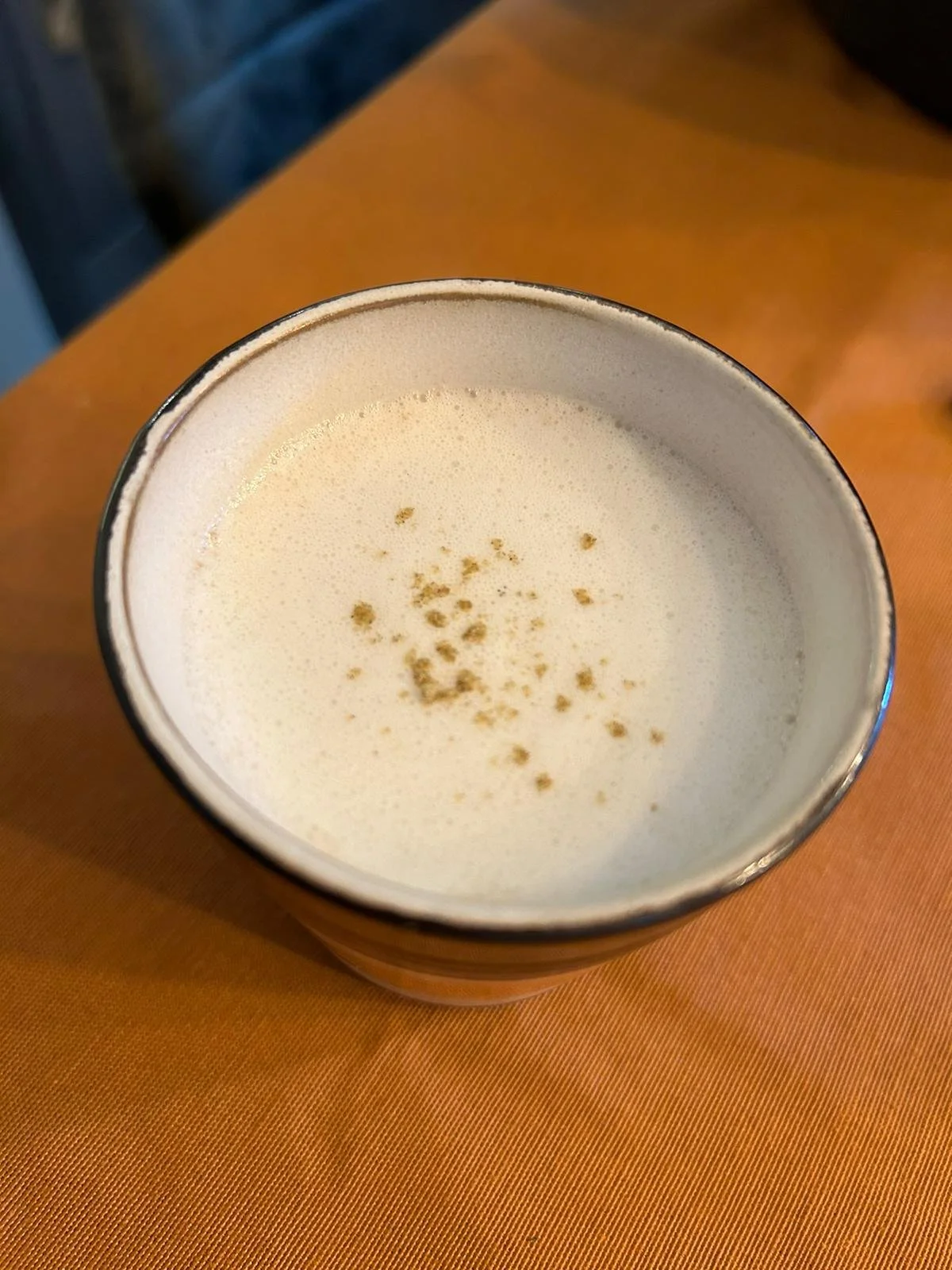 Creamy beverage with foam and sprinkled cinnamon on top, in a ceramic cup, resting on an orange surface.