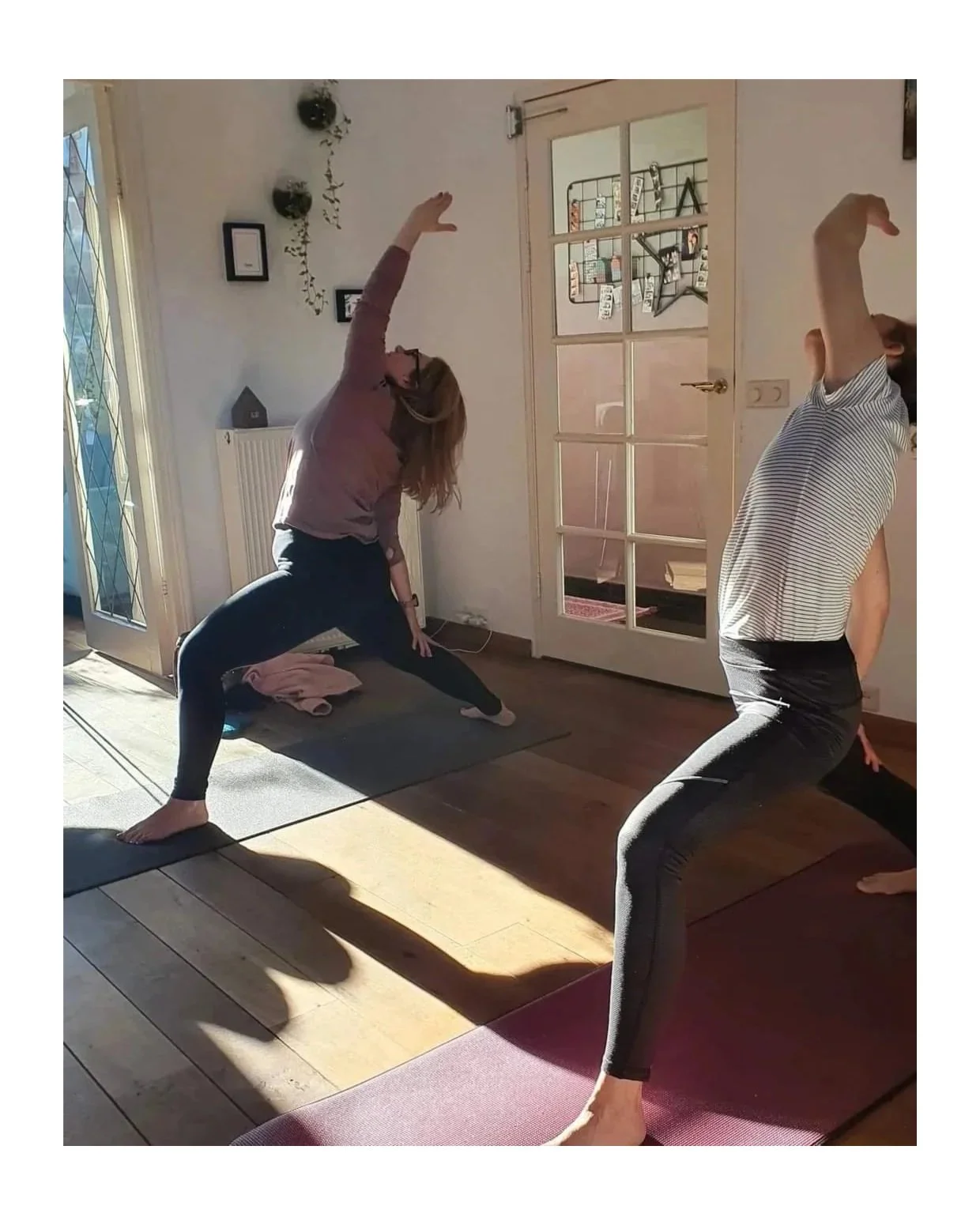 Two women practicing yoga indoors, in a lunge position with arms extended overhead, on yoga mats in a sunlit room.