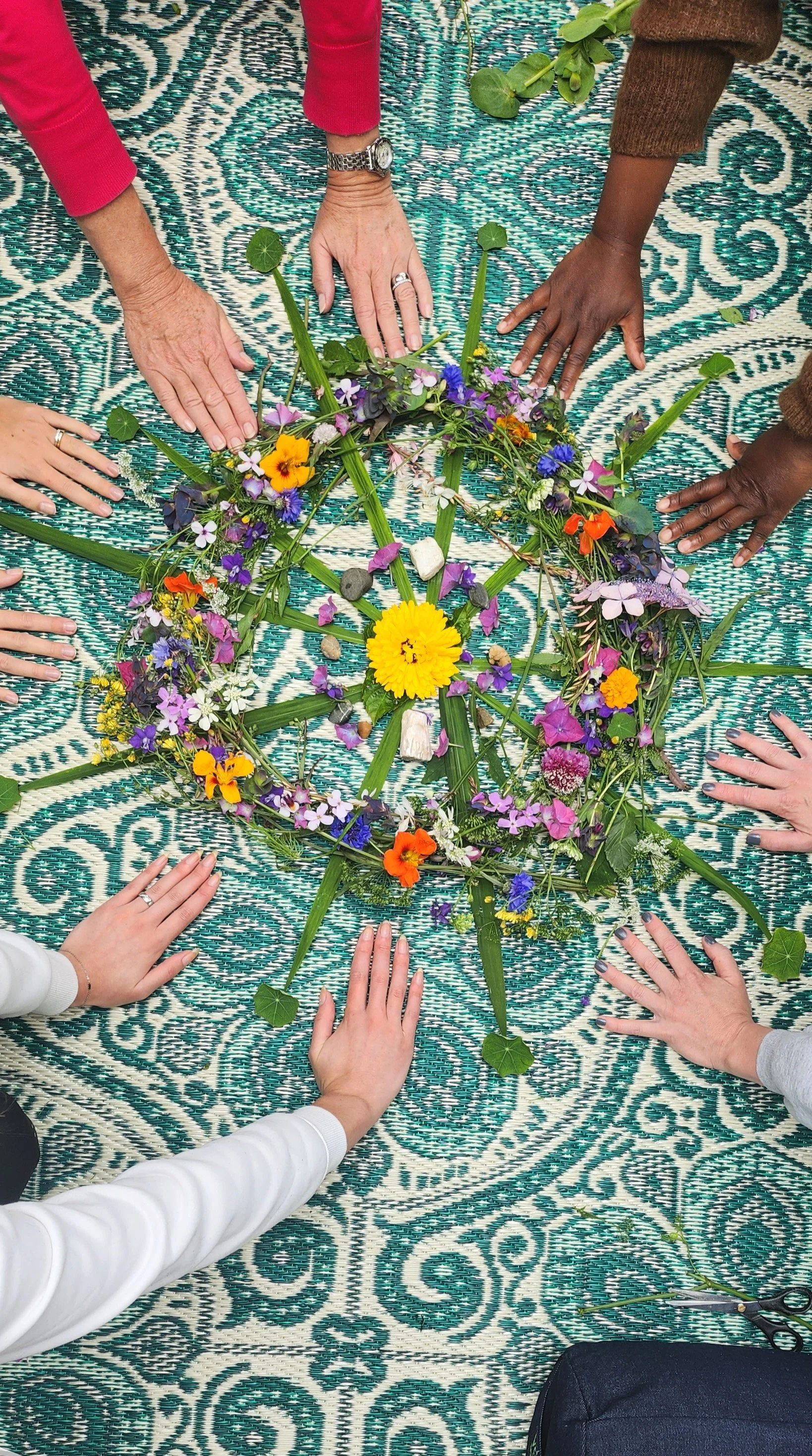 A group of hands placed around a circular floral arrangement with colorful flowers on a patterned teal and white mat.