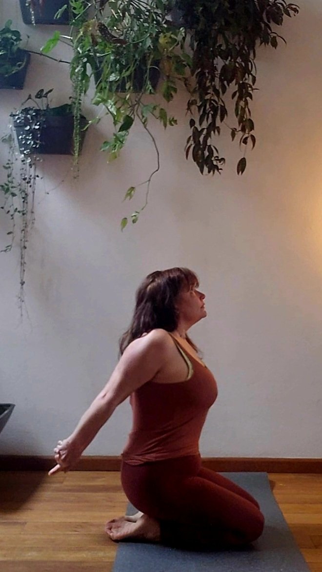 A woman practicing yoga indoors on a mat, in a kneeling position with her arms extended behind her and hands clasped, near a wall with hanging plants.