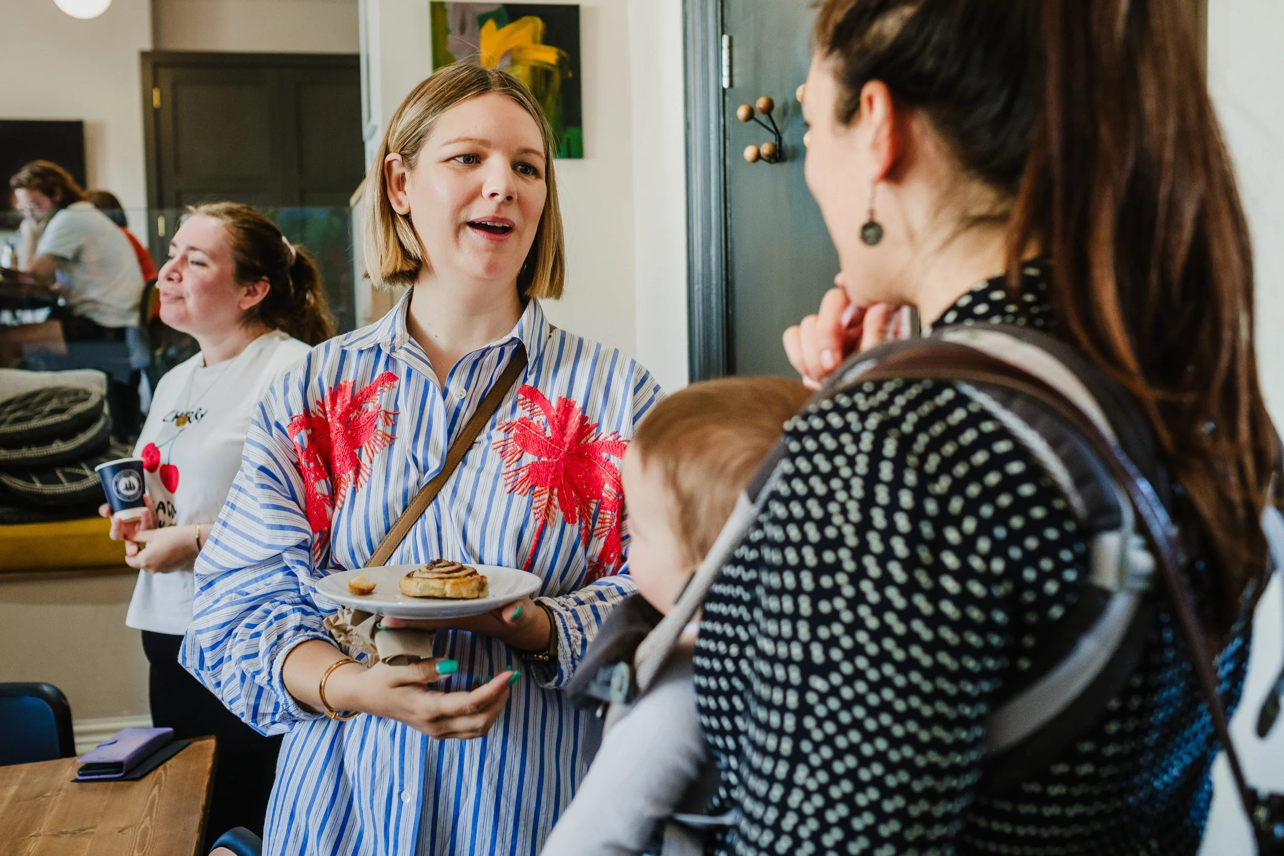 A woman in a blue striped shirt with red embroidery on the shoulders is holding a plate with a cinnamon roll and engaging in conversation with a woman carrying a baby in a black and white polka dot shirt, in a cafe or restaurant setting.