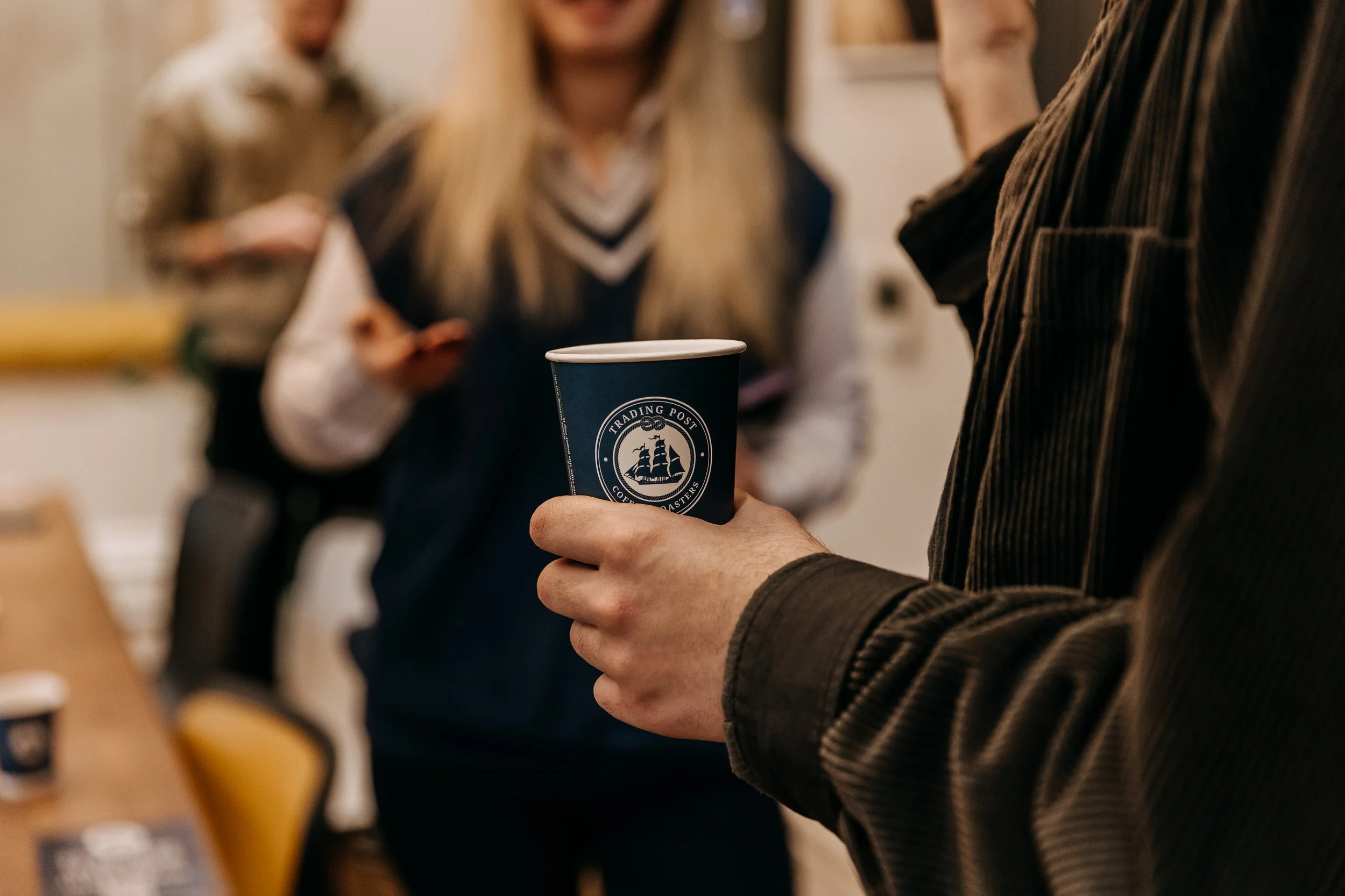 Close-up of a person holding a blue paper coffee cup with a logo, in a social setting with other people in the blurred background.