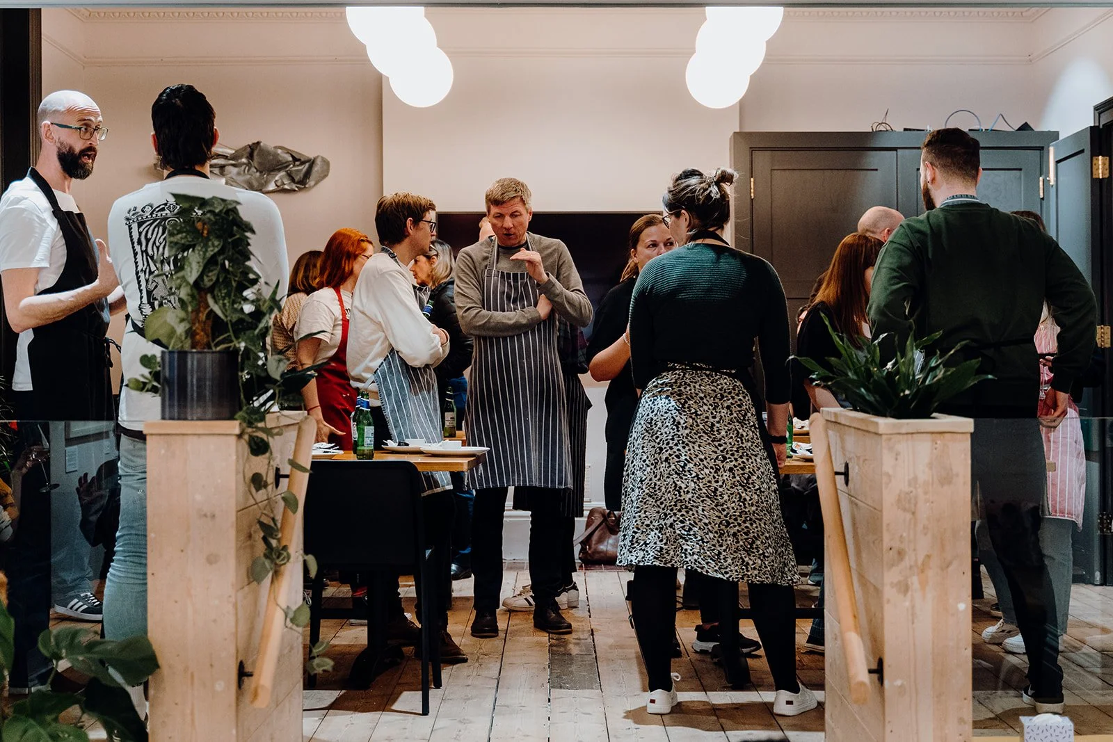 Group of people gathered in a cooking class, wearing aprons, in a kitchen with plants and warm lighting.