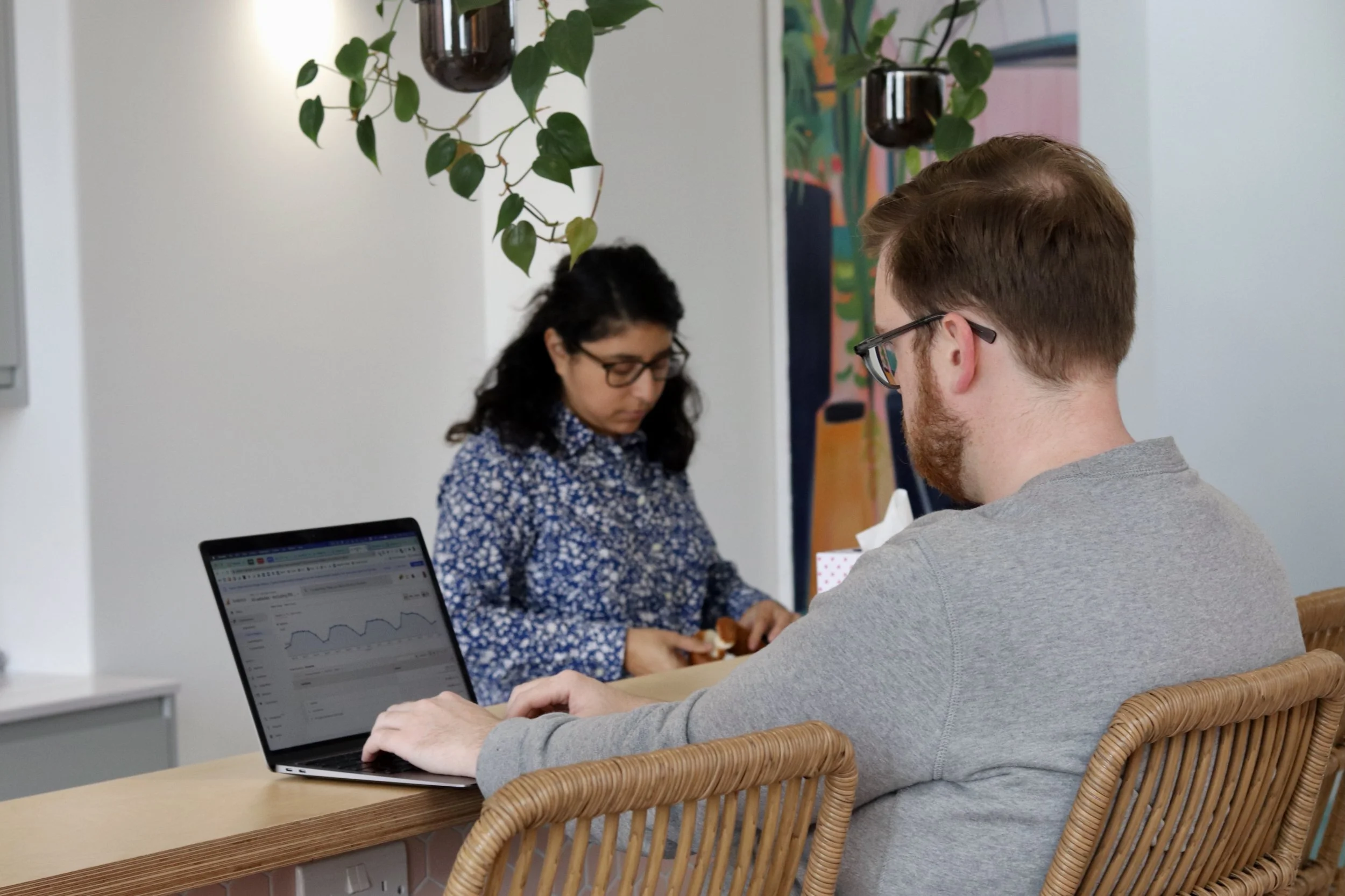 A man with glasses working on a laptop at a wooden table, while a woman with glasses looks down and searches in a small bag.