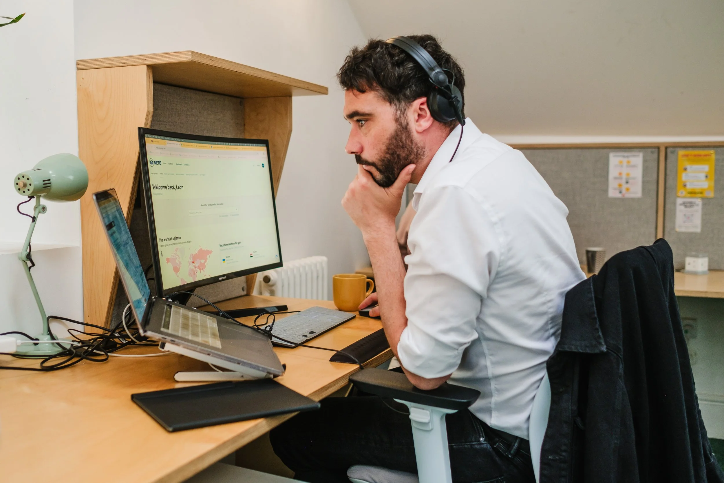 Man with dark hair and beard wearing headphones, white shirt, and black pants, sitting at a cluttered desk with multiple screens, looking at the computer screen with a focused expression.