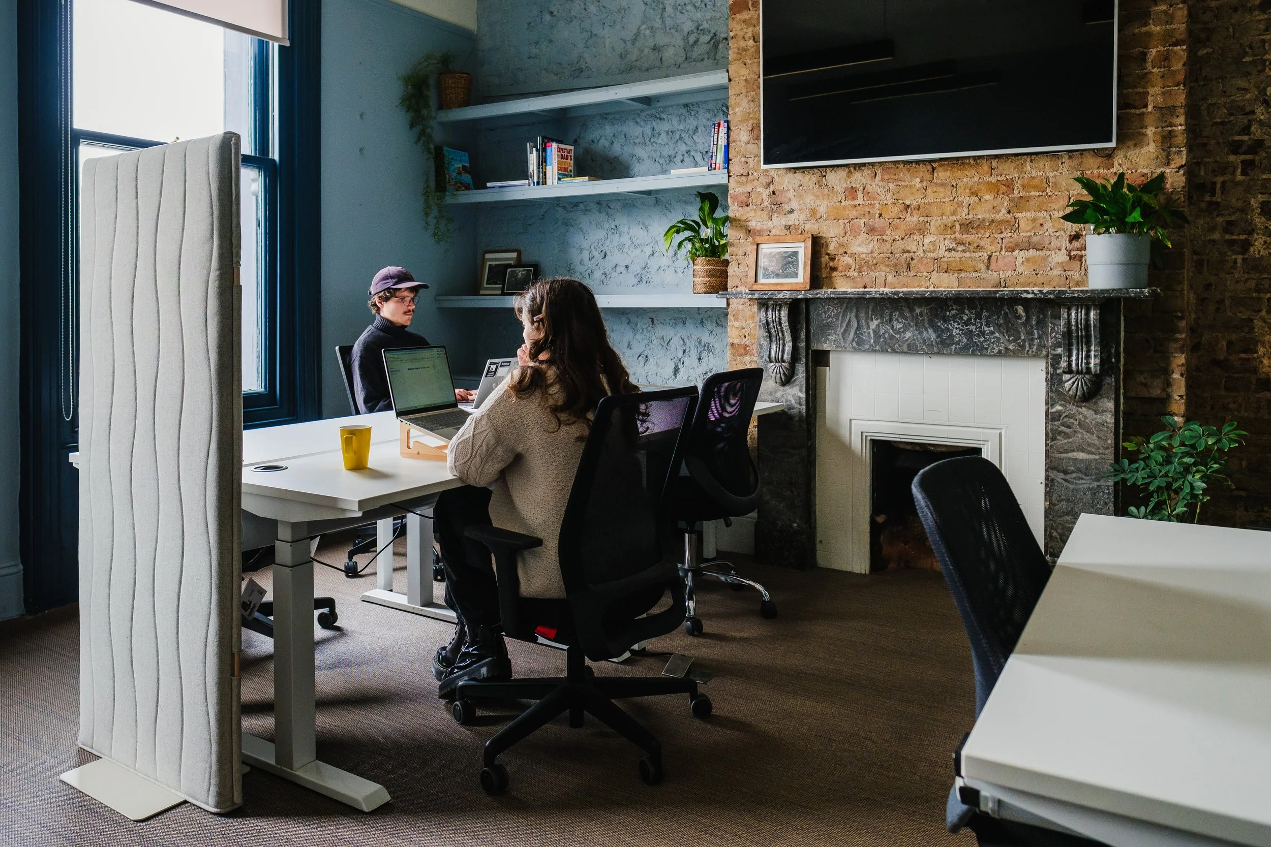 Two people working at a shared desk with computers, books, and office supplies in an office with exposed brick wall and colorful abstract paintings.