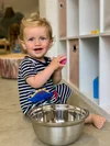 A smiling toddler with blue eyes and blonde hair, wearing a navy and white striped shirt with colorful dinosaur patches, sits on the floor holding a pink toy while next to a metal mixing bowl.