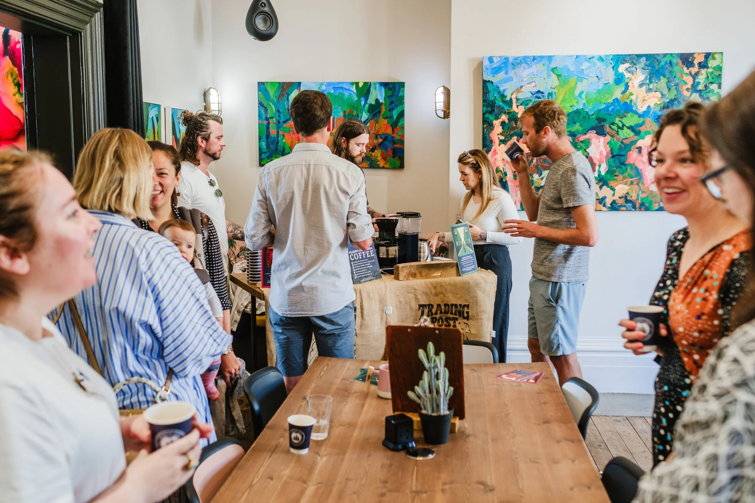 People gathered inside a coffee shop, some ordering at the counter and others holding coffee cups, with colorful abstract paintings on the walls.