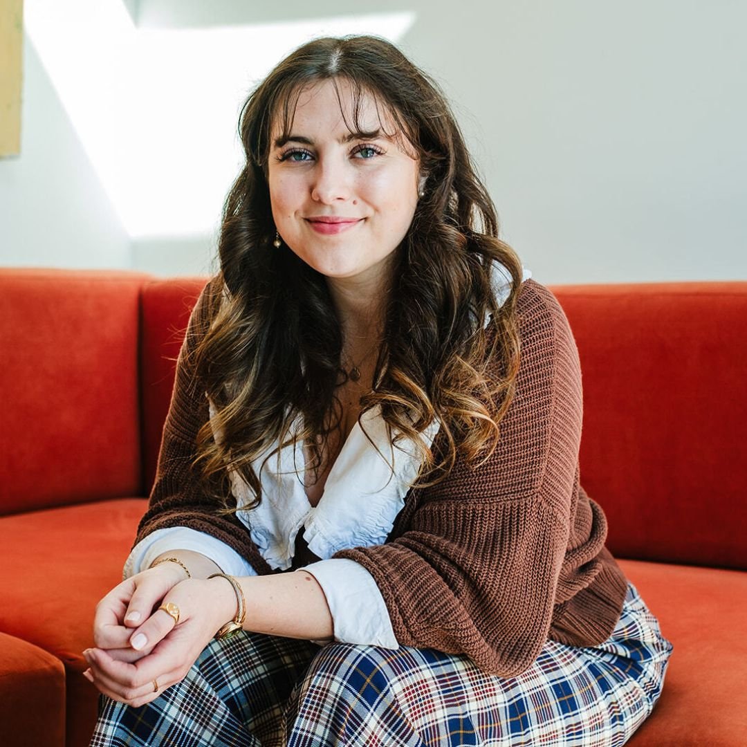A young woman with long curly brown hair, wearing a brown cardigan with a white collar and checkered trousers, sitting on a red sofa with her hands folded in front of her with gold rings and a gold watch.