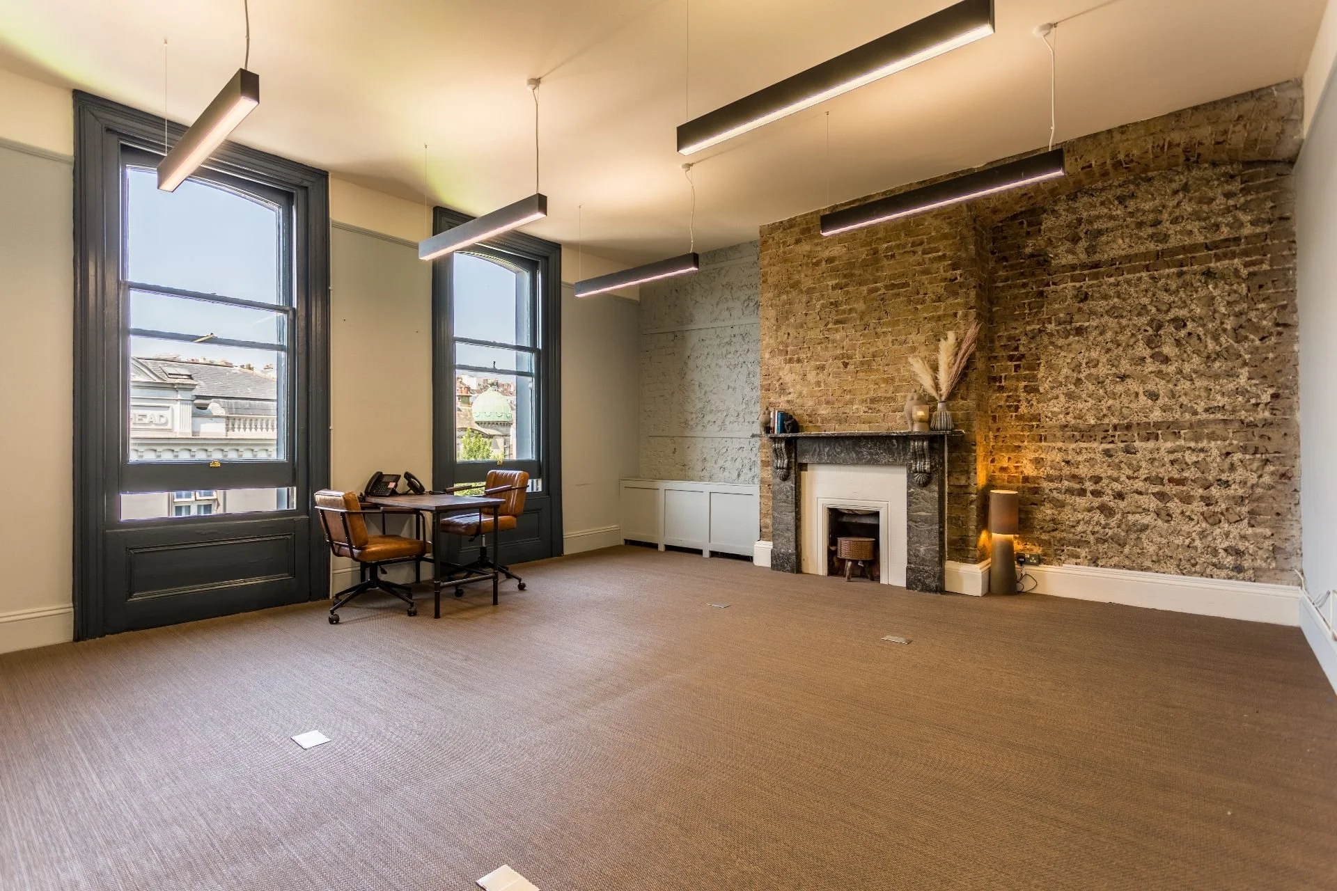 Empty room with large windows, brown carpet, and a brick fireplace with a dark marble mantle. There is a small table with two brown chairs near the windows, and decorative items on the fireplace mantel. Modern lighting fixtures hang from the ceiling.