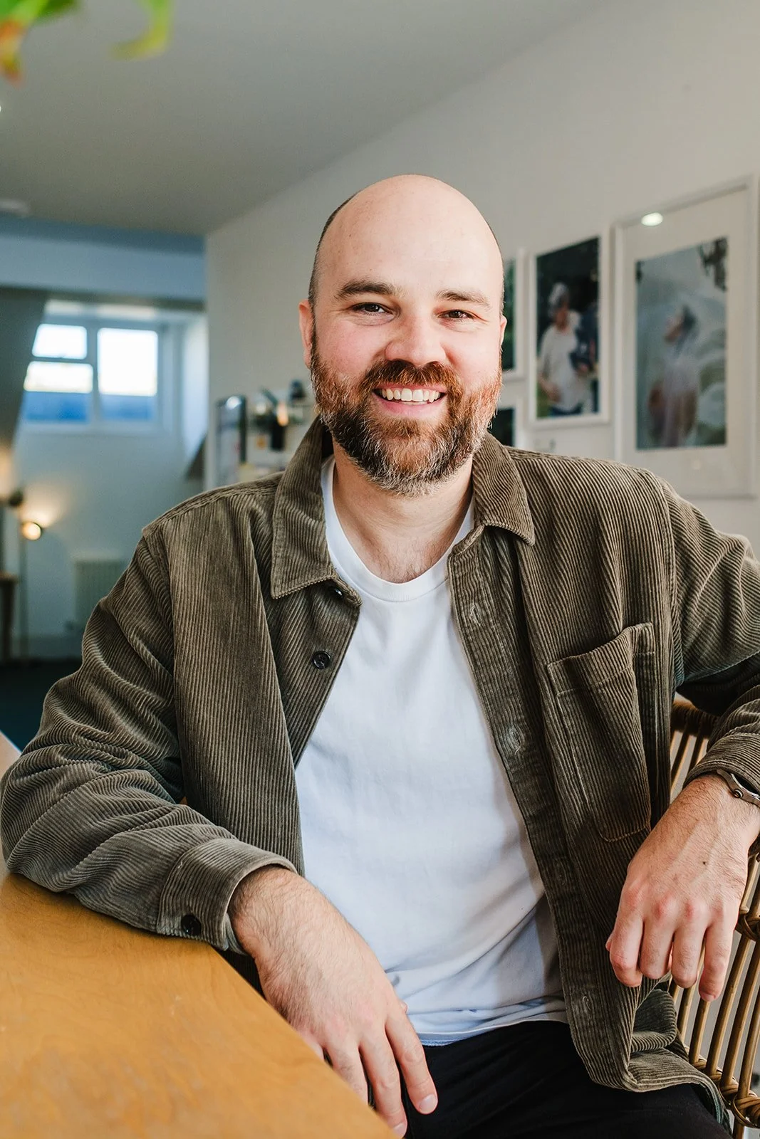 A man with a beard and shaved head smiling, sitting at a table in a cozy, well-lit room with framed pictures on the wall.