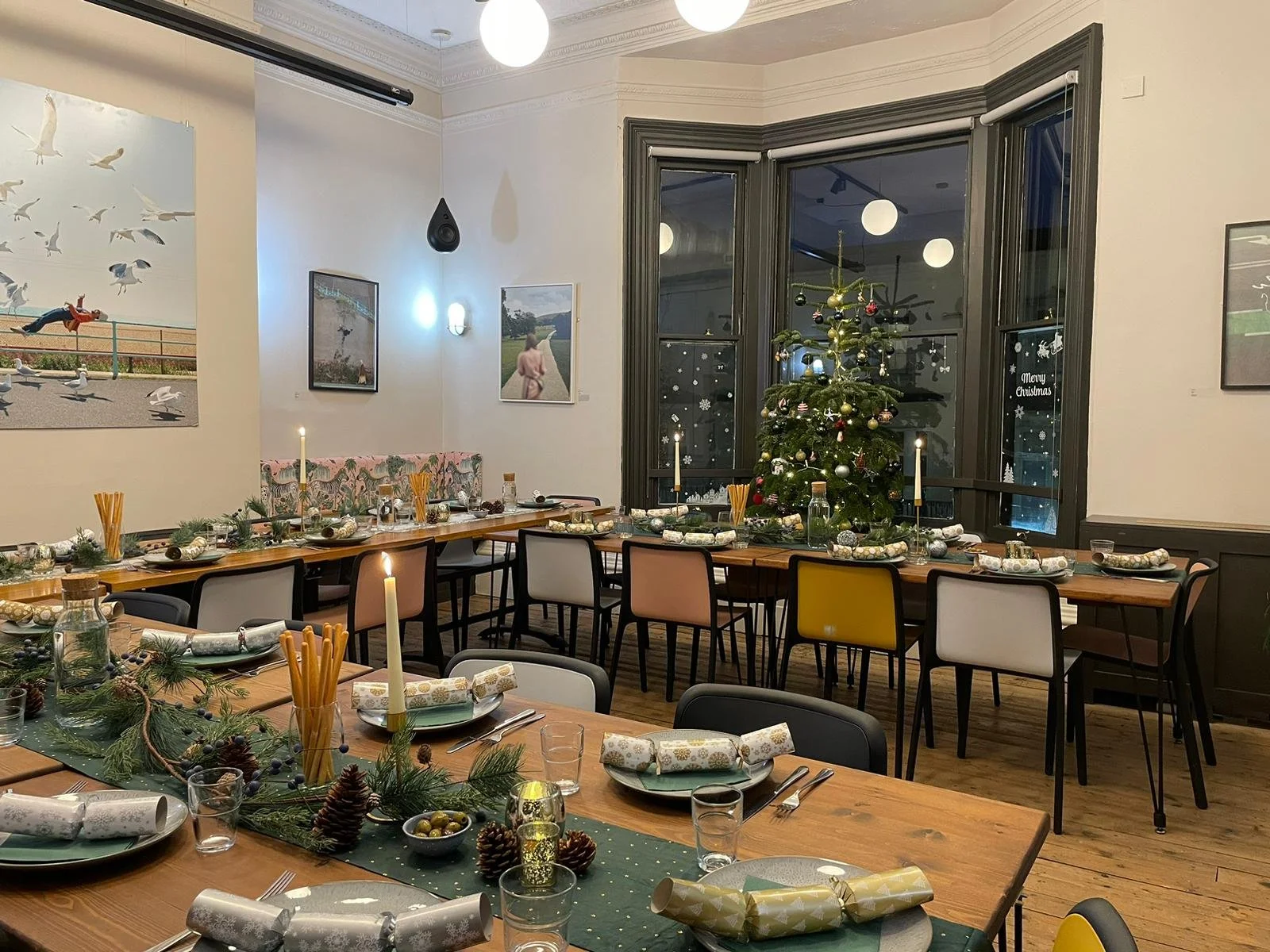 Christmas decorated dining room with a Christmas tree, tables set with holiday-themed decorations, lit candles, and wrapped presents, in front of large bay windows.