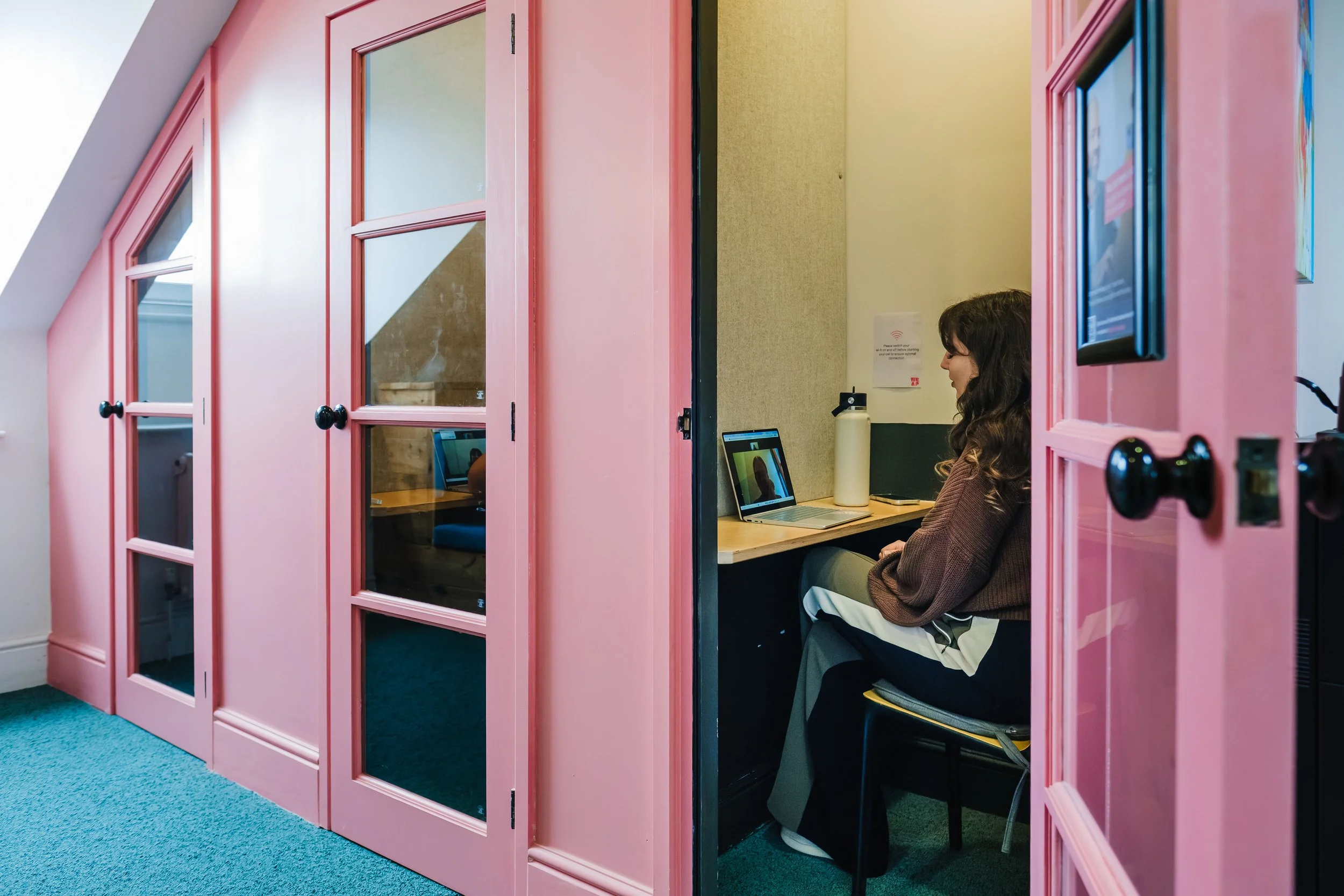 3 pink phone booths with a woman sat at the one on the right on a video call 