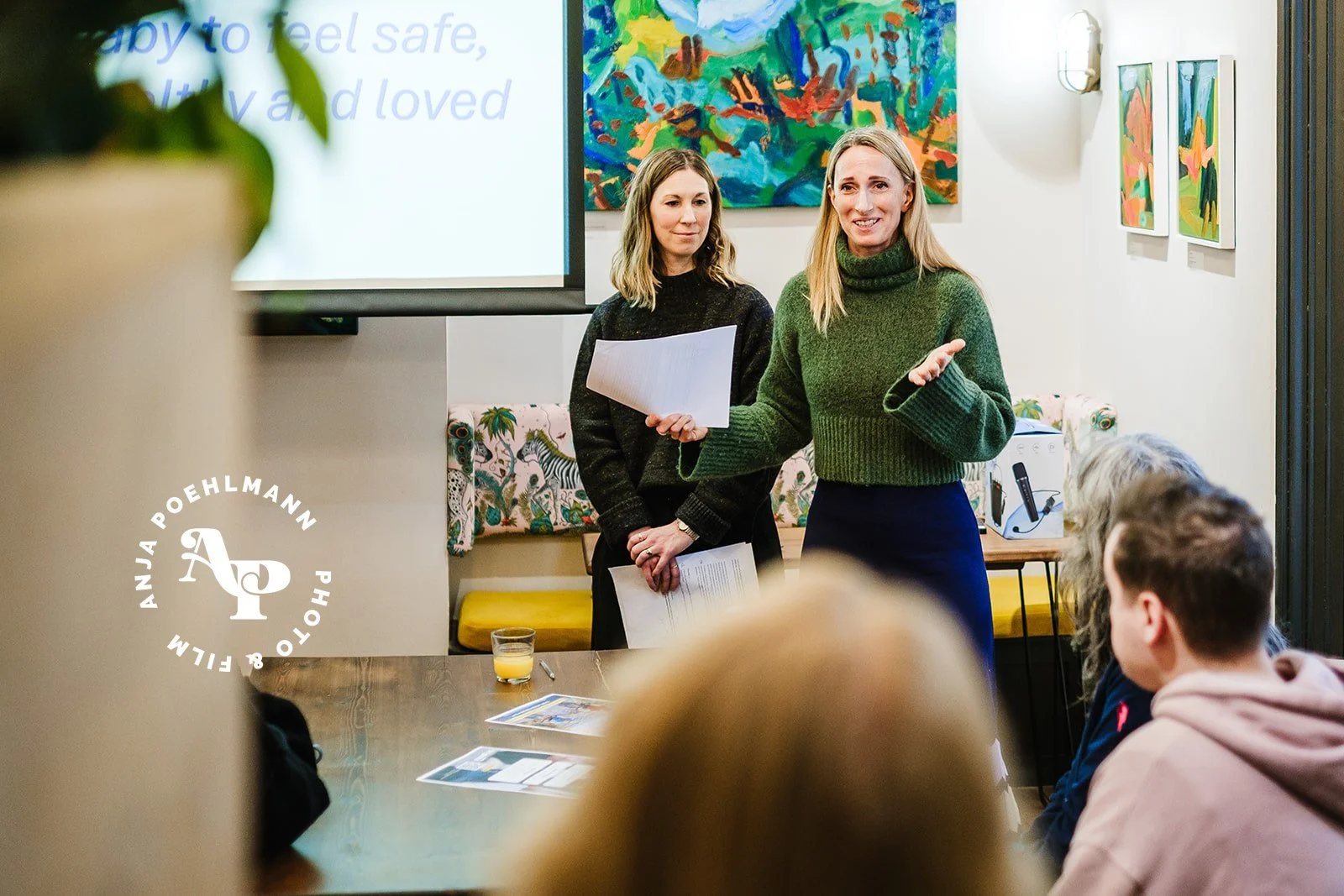 Three women and a baby on the beach near Brighton Palace Pier. One woman is holding the baby in a baby carrier, while the other two women are looking at a tablet and a bag. The pier sign is visible in the background.
