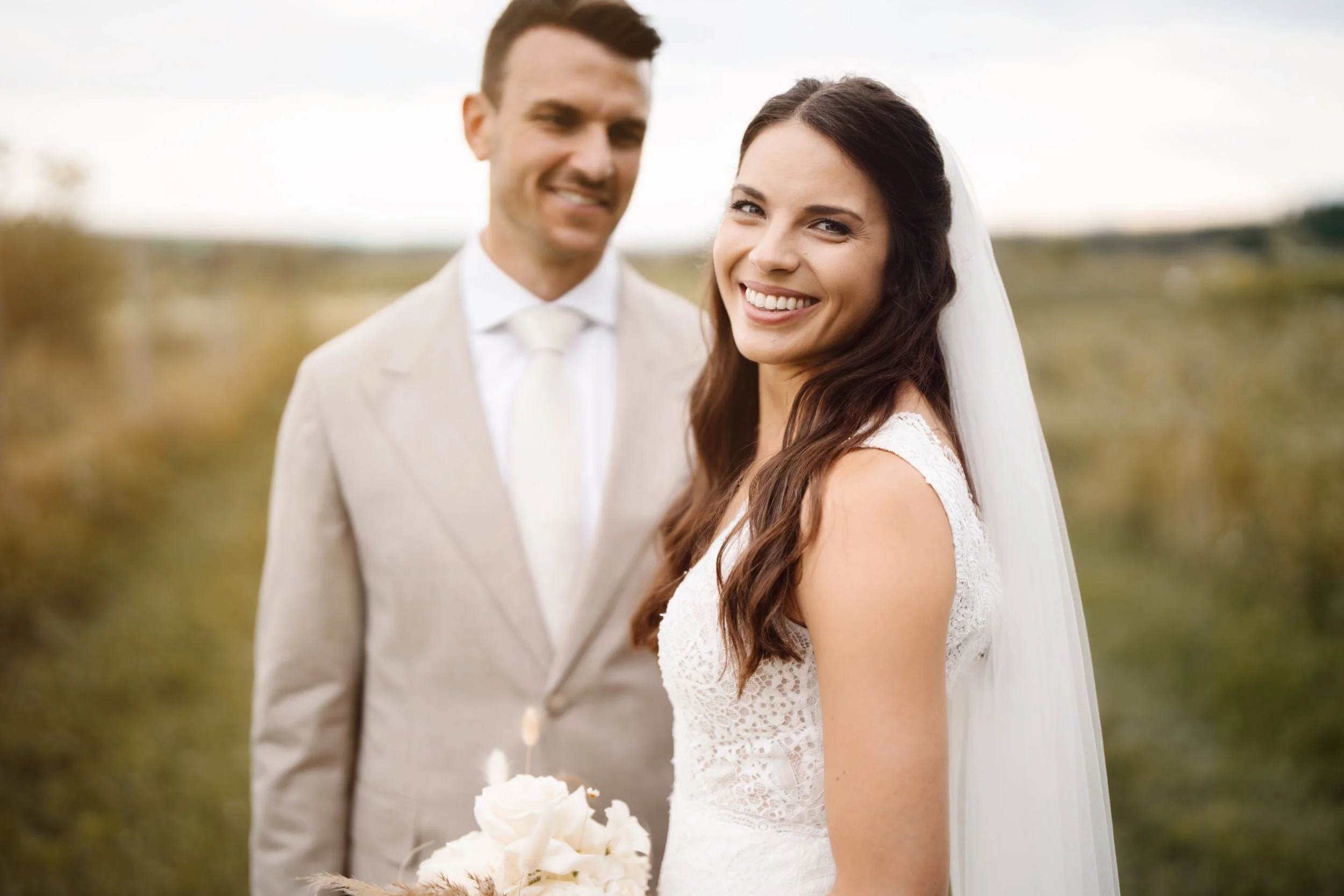 Ein glückliches Brautpaar im Hochzeitskleid und Anzug steht im Freien auf einer Wiese, die Frau hält einen Strauß Blumen.