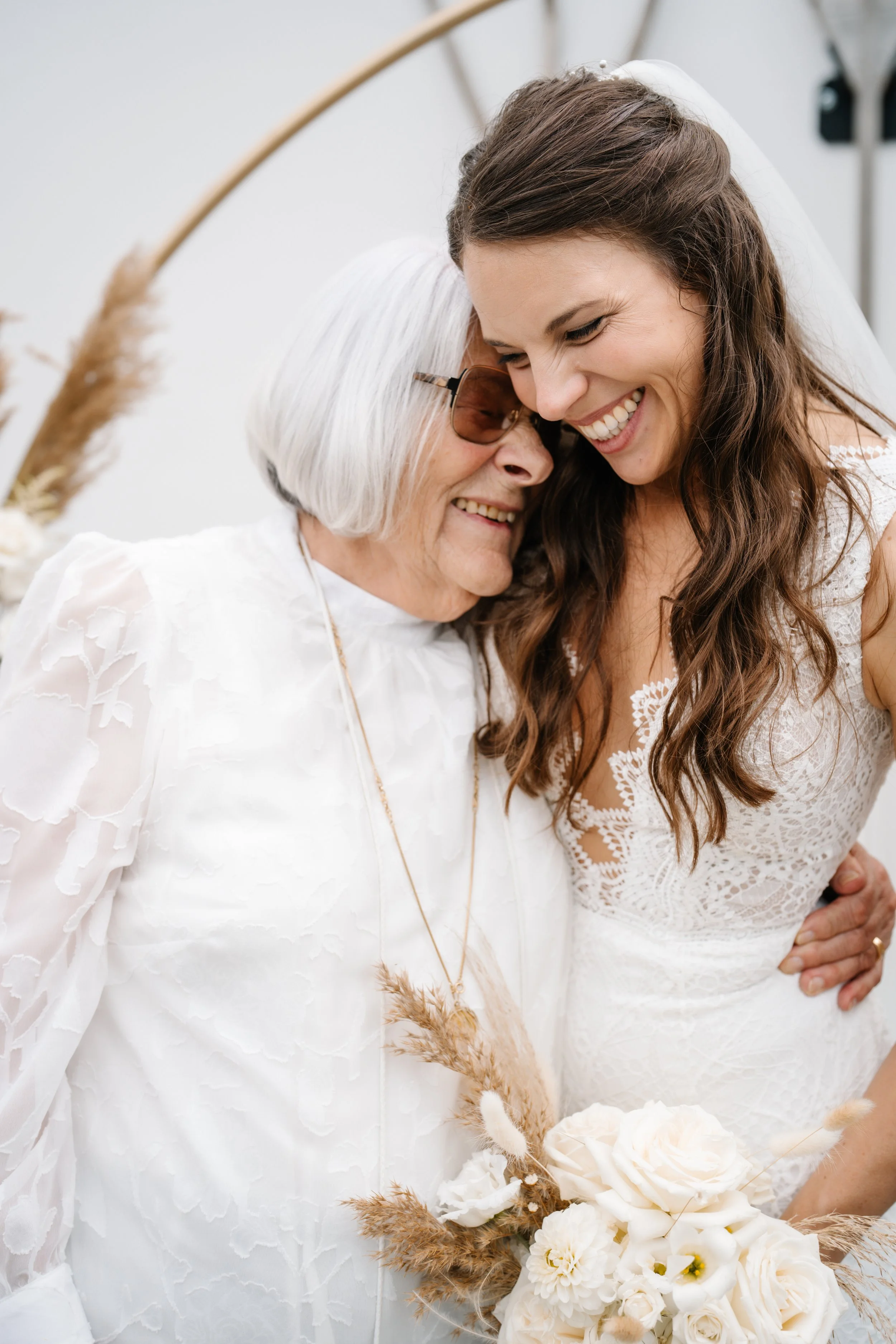 Zwei Frauen, eine ältere mit weißen Haaren und Sonnenbrille, und eine jüngere in einem weißen Brautkleid, umarmen sich glücklich bei einer Hochzeitsfeier.