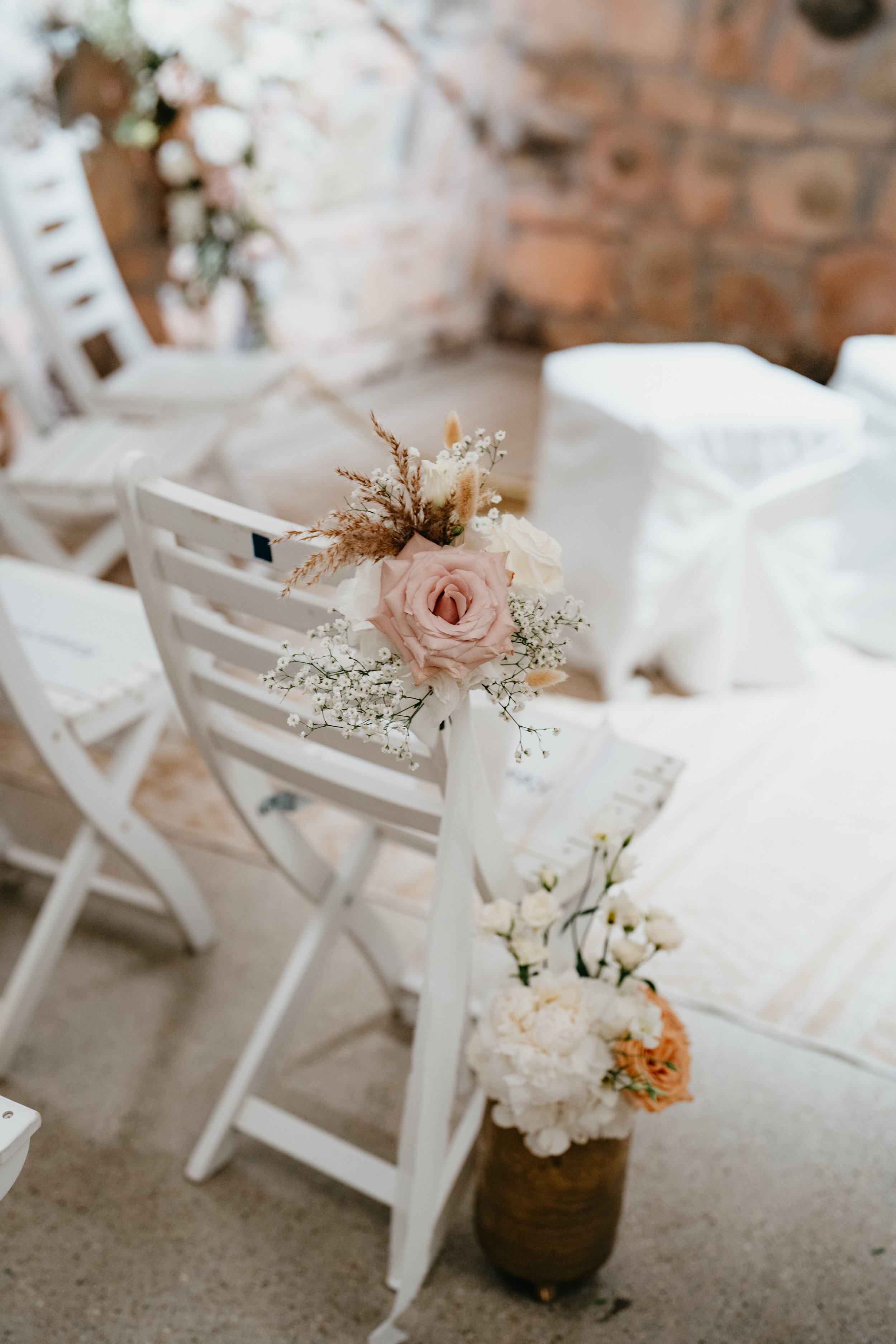 Blumenarrangement an einem weißen Stuhl, bestehend aus einer großen rosa Rose, weißen Blumen und getrocknetem Gras, neben einem Vasen mit weiteren Blumen in einem Raum mit Steinmauer.