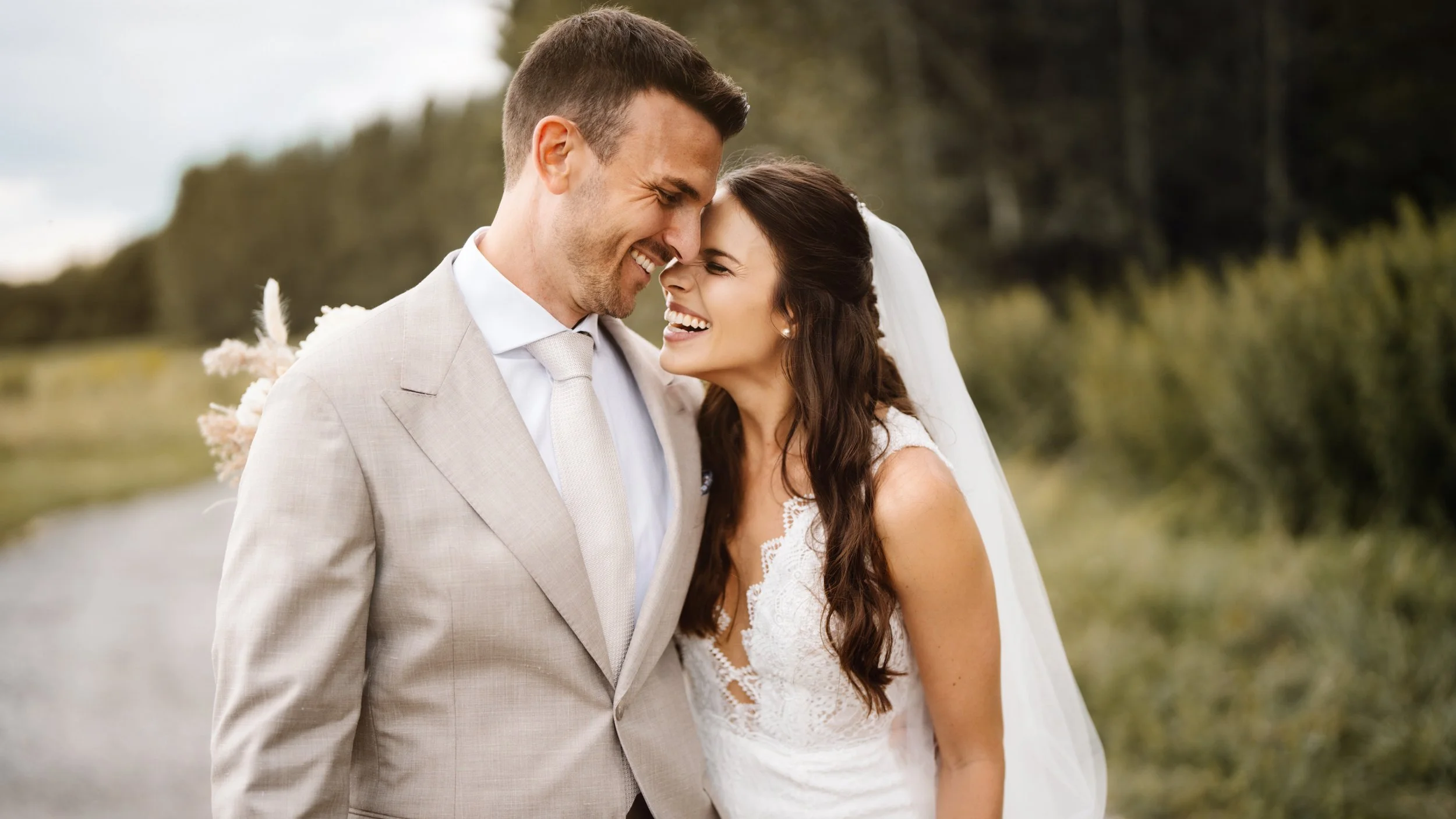 Ein glückliches Brautpaar steht draußen, lacht und berührt ihre Stirn. Die Frau trägt ein weißes Hochzeitskleid mit Spitzenmuster, der Mann trägt einen hellgrauen Anzug mit Krawatte.