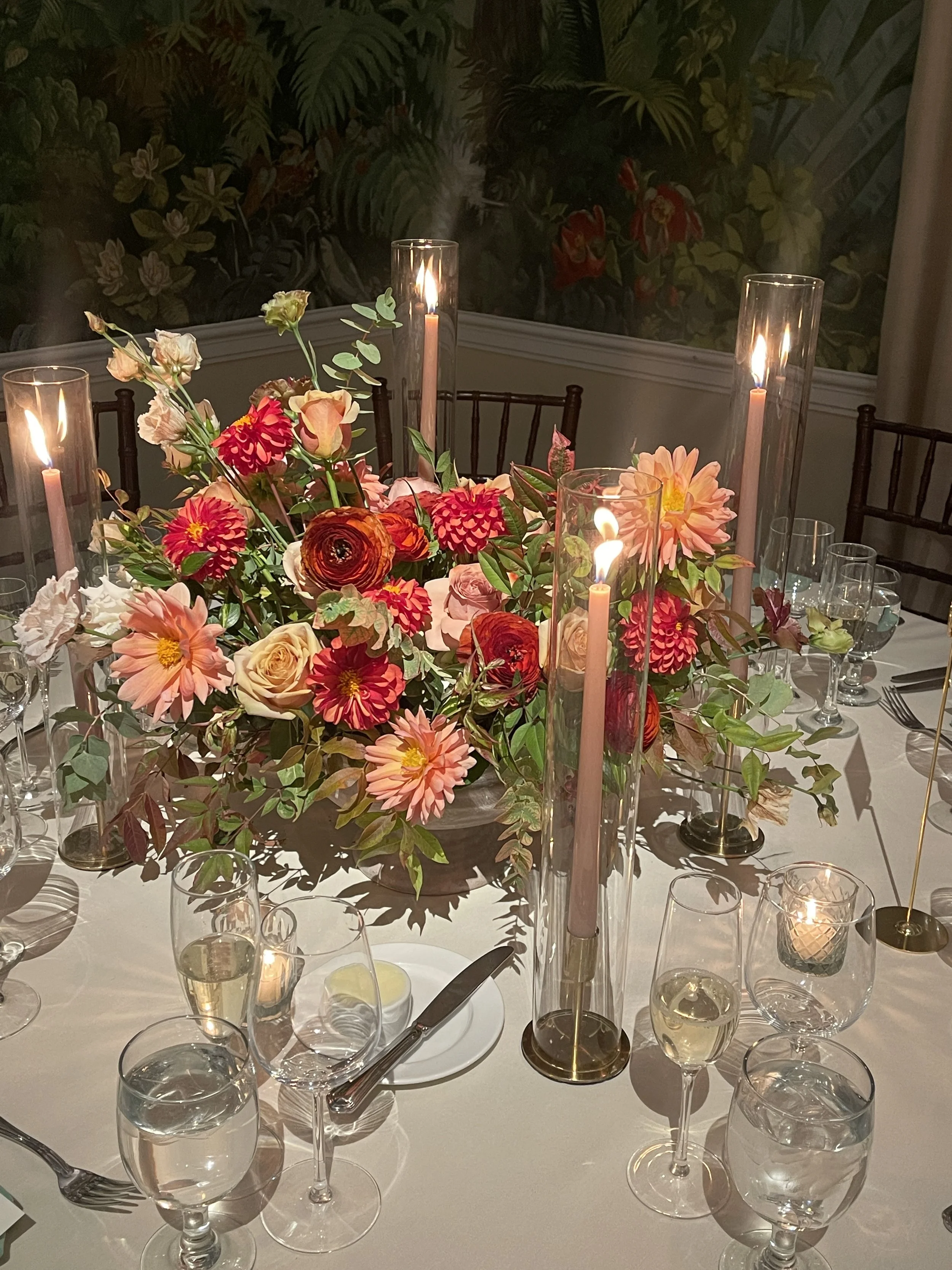 Elegant dining table with centerpiece of pink and red flowers and tall candles, set with wine glasses and cutlery for an event.