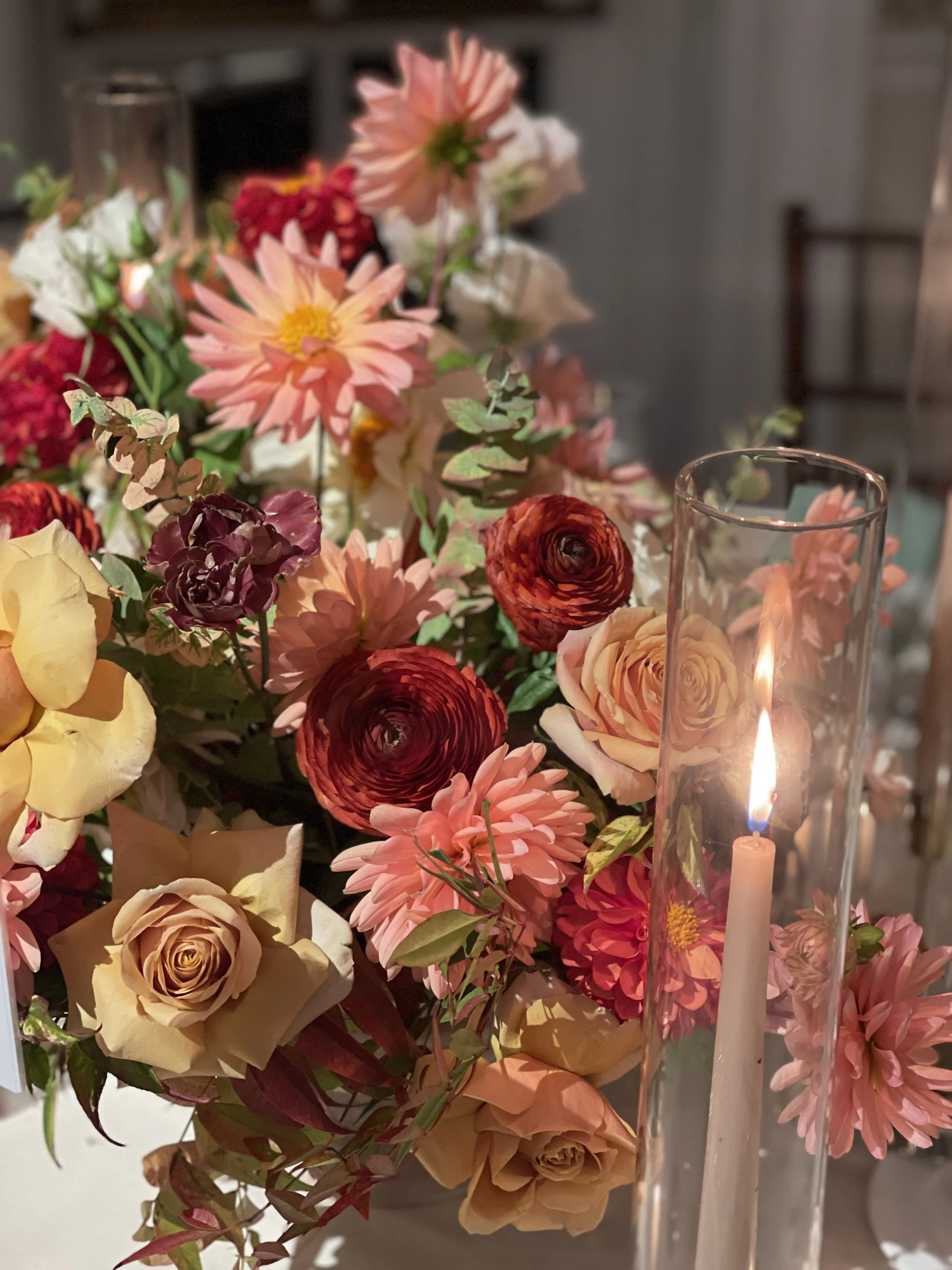 A floral arrangement with pink, peach, yellow, red, and burgundy flowers accompanied by a lit candle in a glass holder.