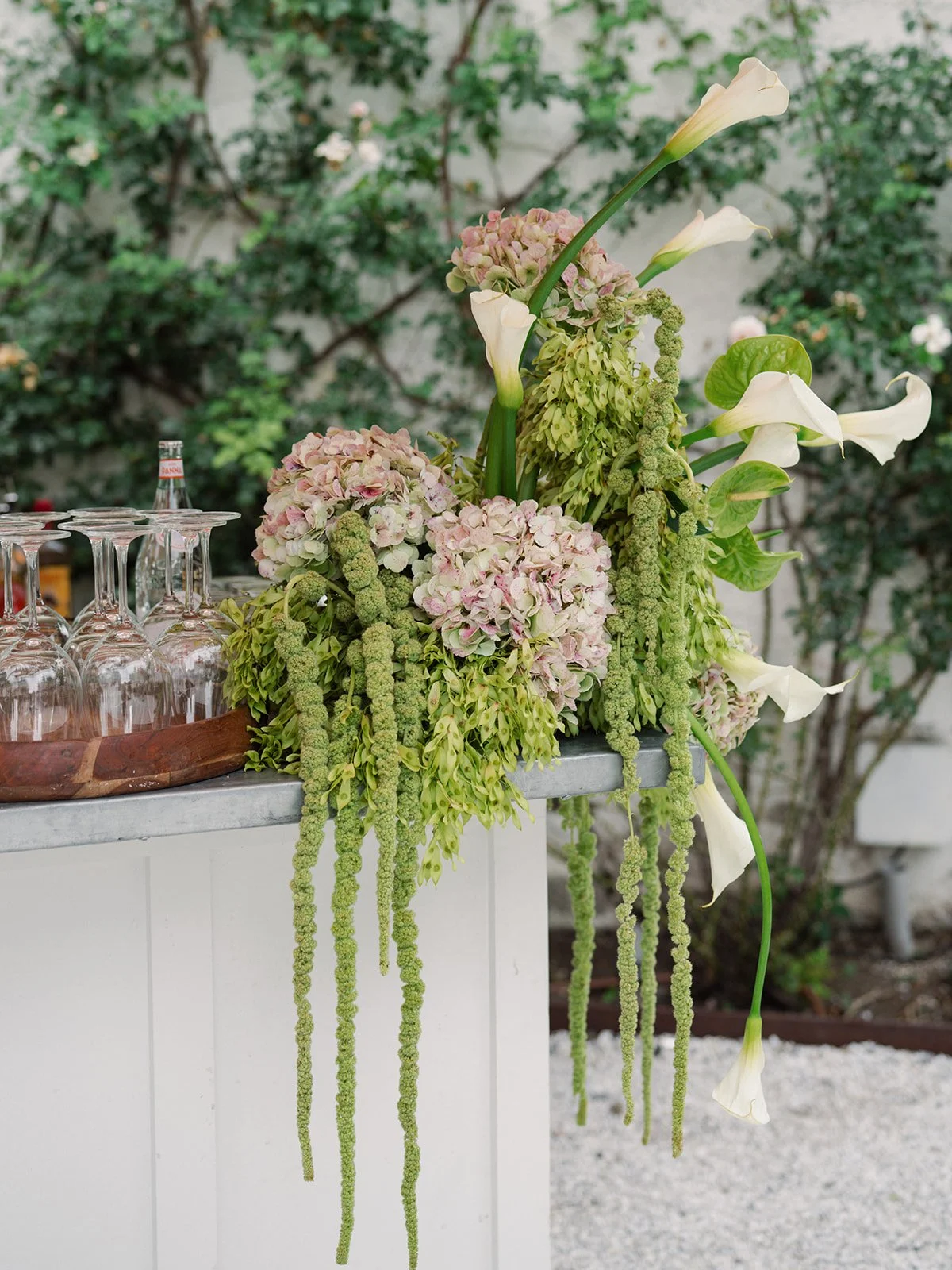 A floral arrangement with white calla lilies, pink hydrangeas, green amaranthus, and other greenery on a white surface outdoors.