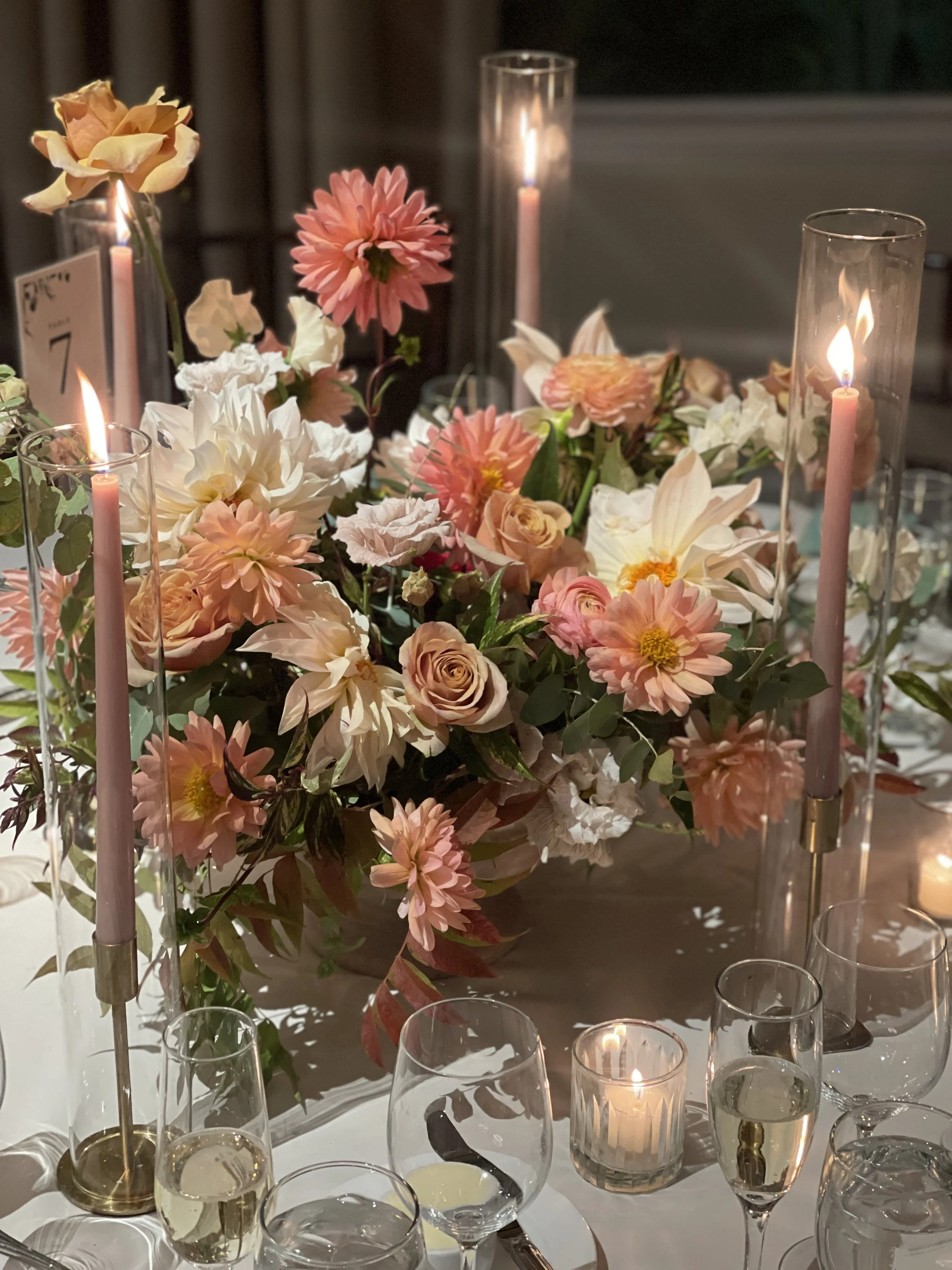 Elegant table centerpiece with a large bouquet of pink, peach, white, and cream flowers, surrounded by tall glass candle holders with pink taper candles and lit votive candles, set on a white tablecloth.