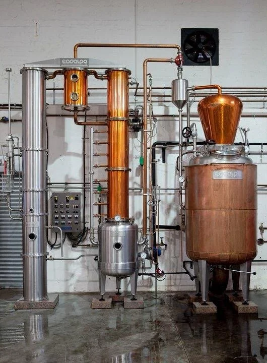 Copper and stainless steel distillation equipment in a distillery, including a condenser, boiler, and various pipes and gauges.