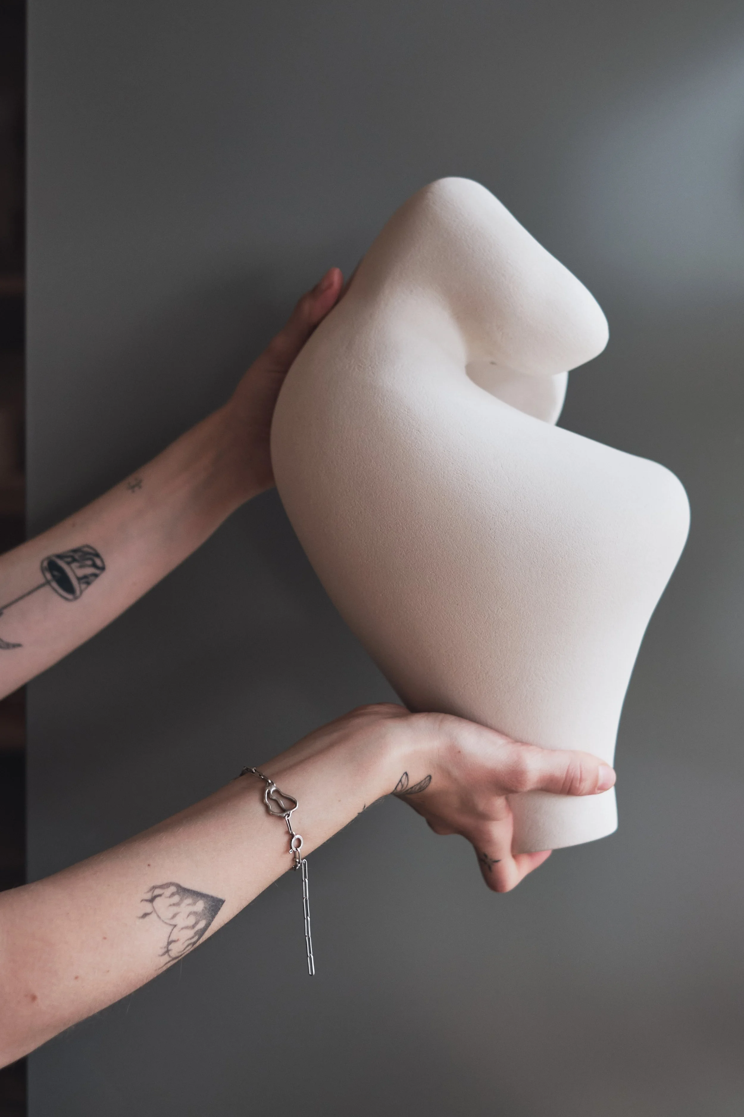 Eine Person hält eine große, weiße, skulpturartige Figur an den Beinen, mit Tattoos an den Armen, einem Armband und einem Kettchen, vor einem grauen Hintergrund.