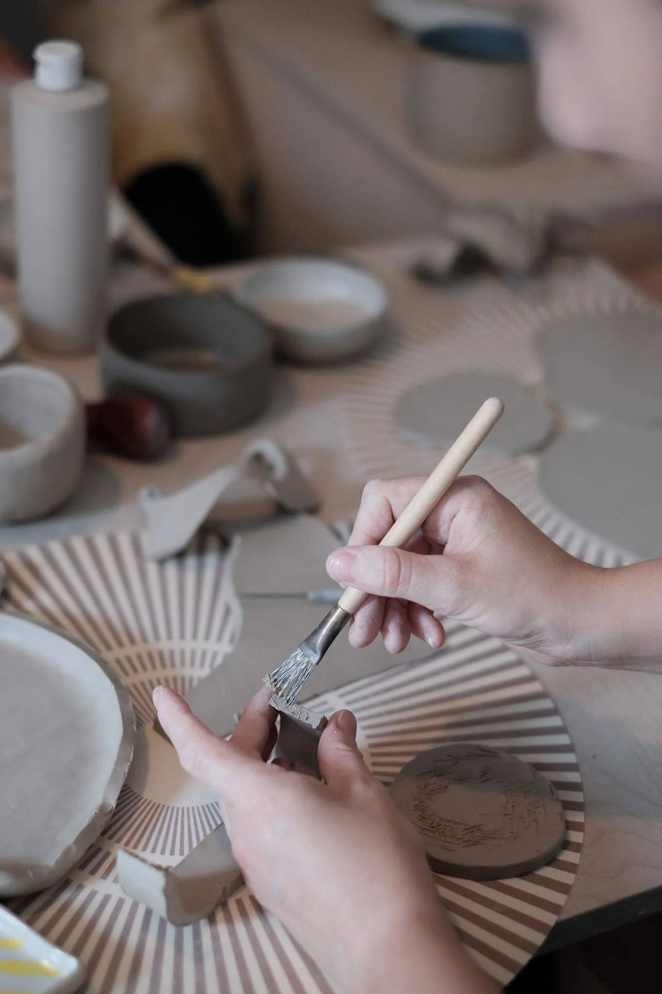 Person working on ceramic pottery, using a brush to glaze or decorate a piece, with various unfinished ceramic pieces and tools on the table.