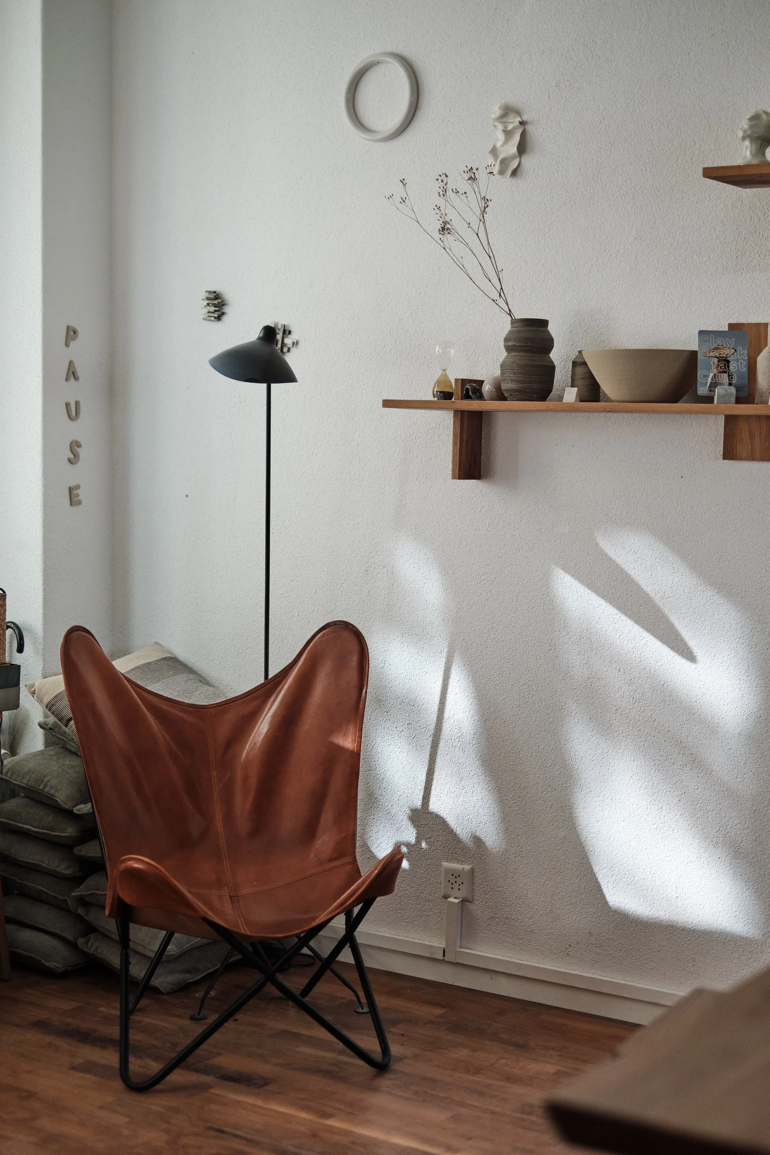 Ein Wohnzimmer mit einem braunen Ledersessel, einem schwarzen Stehlampe, einer Holzregalwand mit Keramikvasen und Dekorationen, einem Bett mit Kissen im Hintergrund, einer weißen Wand, einem Boden aus Holzplanken und einem Stromstecker an der Wand.