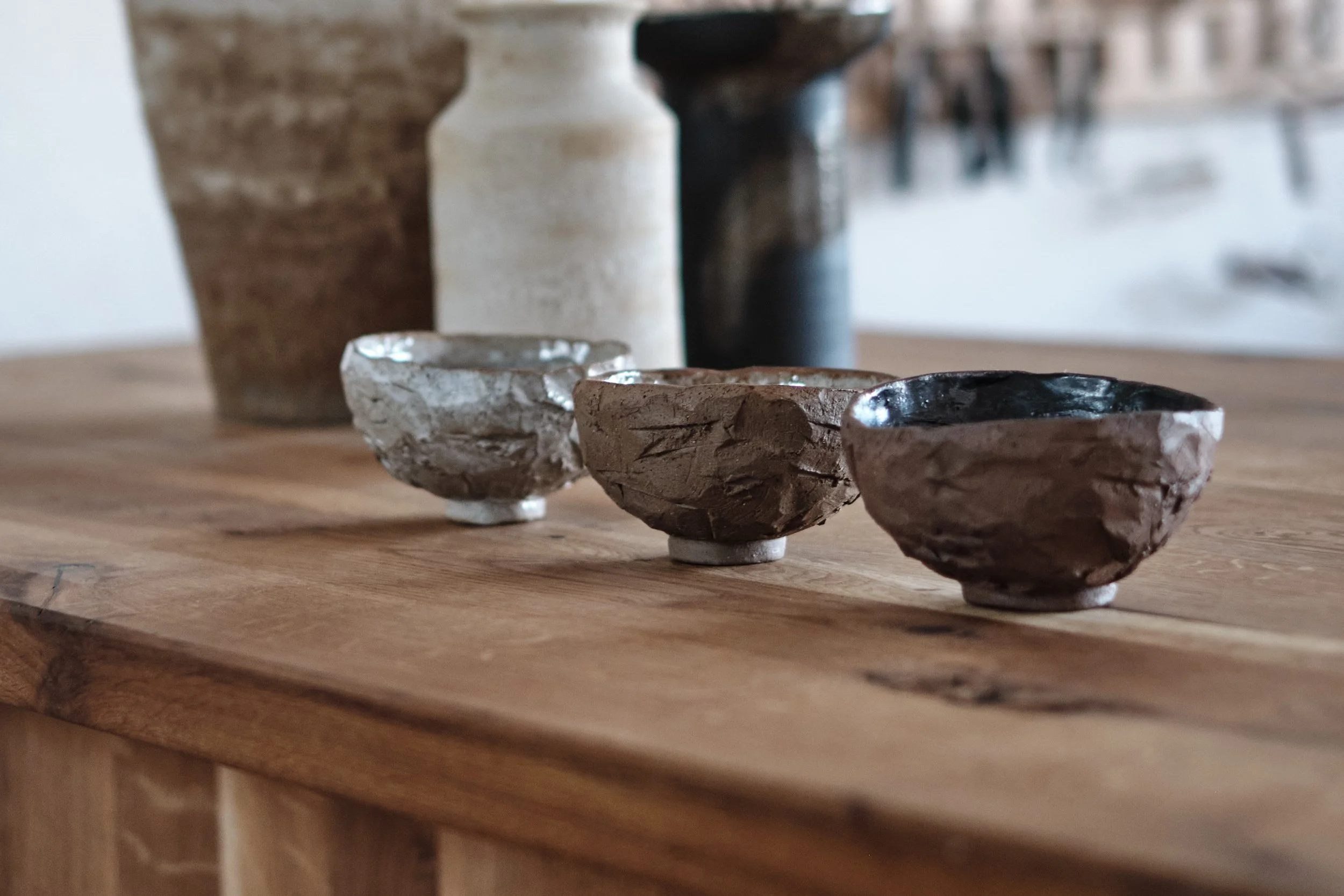 Three rustic ceramic bowls with uneven surfaces and glazed interiors, placed in a row on a wooden surface, with blurred vases and furniture in the background.
