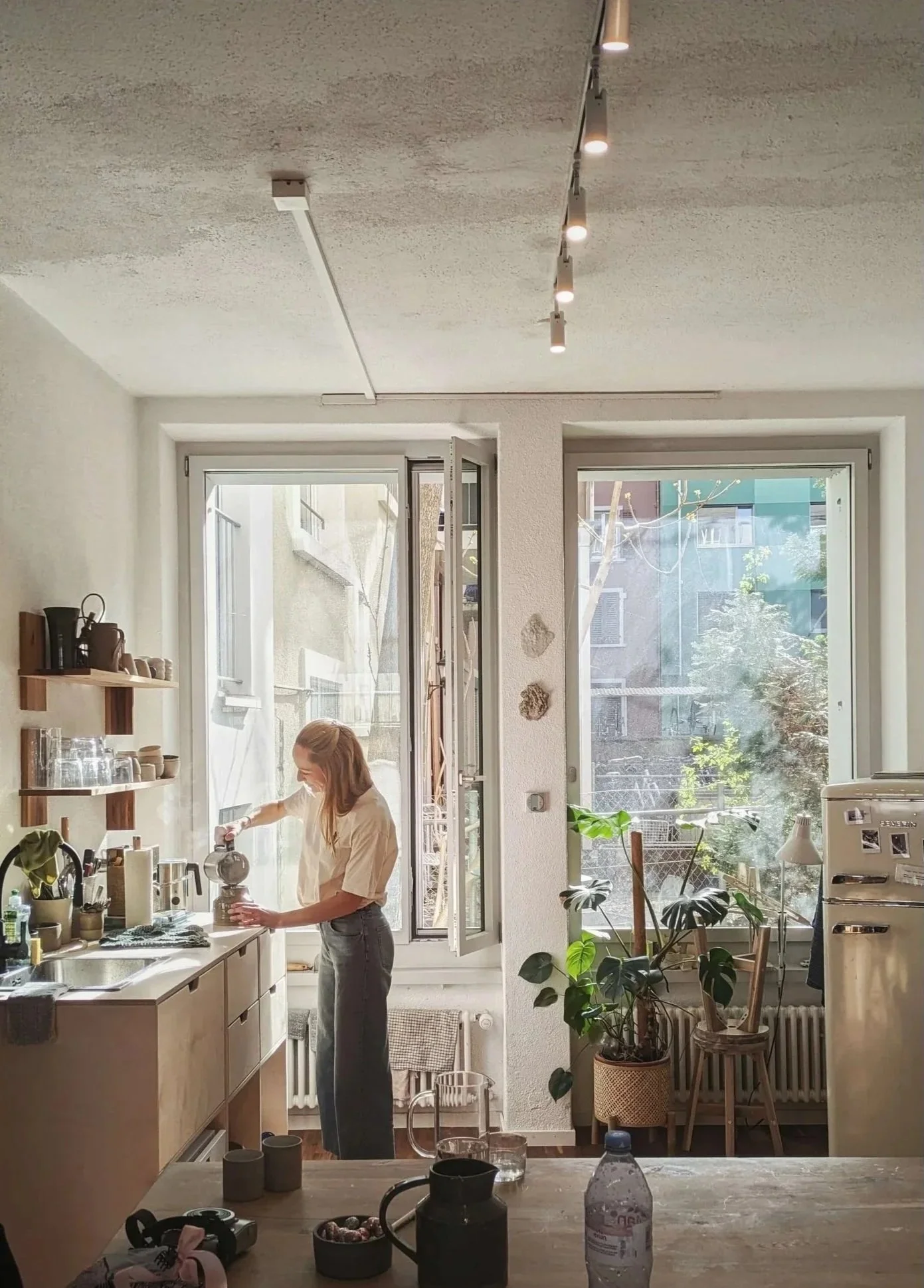 Woman wearing a face mask preparing coffee in a bright kitchen with large front windows, wooden shelves, potted plants, and various kitchen items on a countertop.