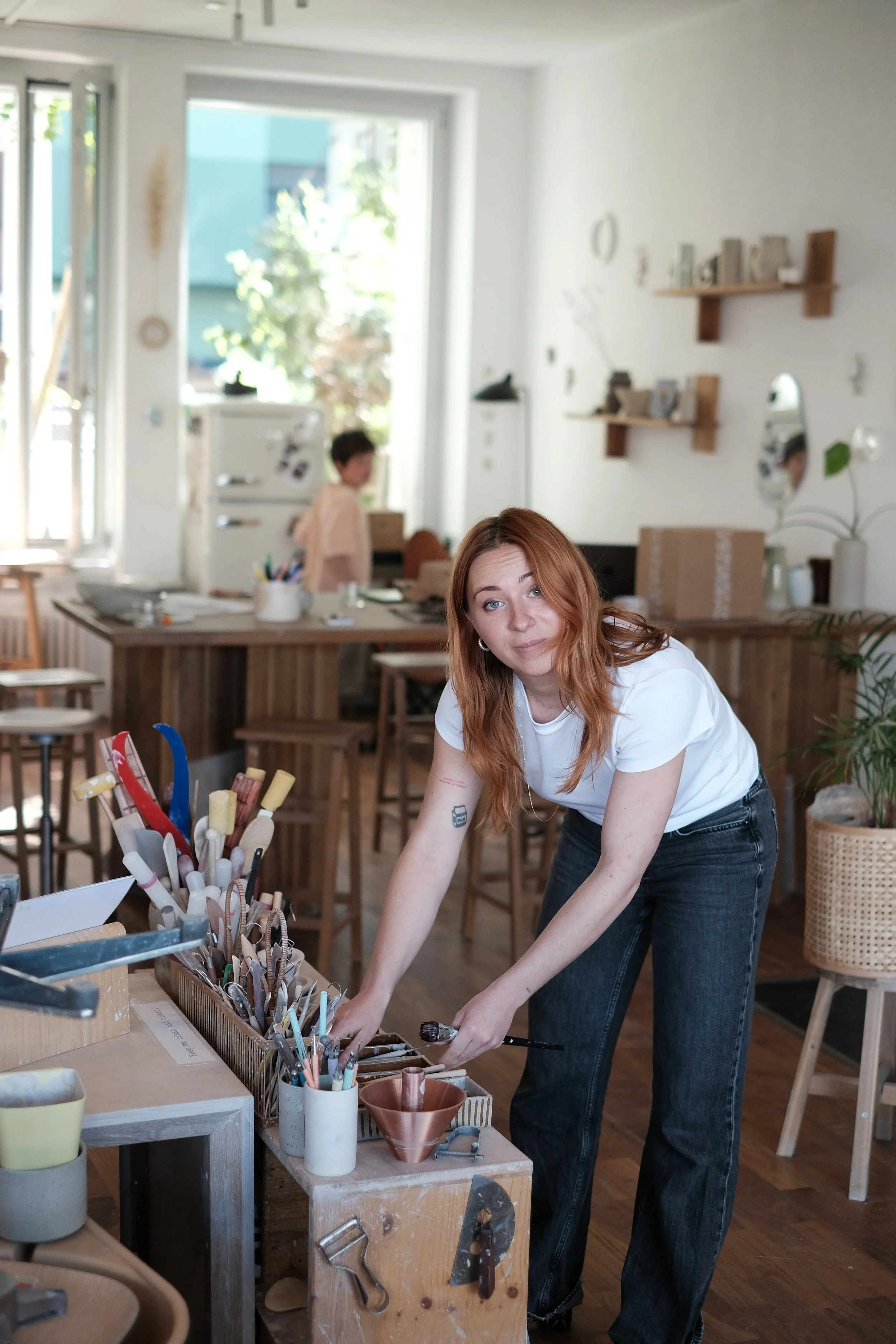 Eine junge Frau mit roten Haaren im Vordergrund, die an einem Tisch mit künstlerischen Materialien arbeitet, im Hintergrund ein Raum mit Regalen und einer blauen Tür, im Hintergrund eine weitere Person, die an einem Tisch sitzt. Anna Team Mitglied